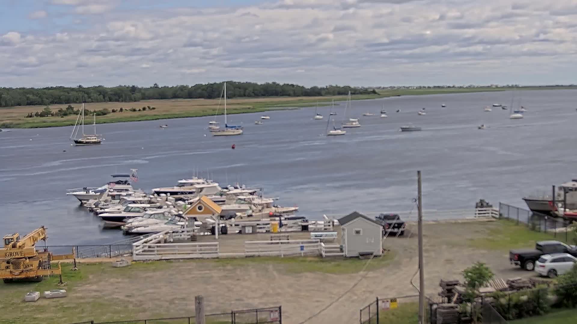 A marina with numerous boats docked, some additional boats are anchored in the water, under a partly cloudy sky.