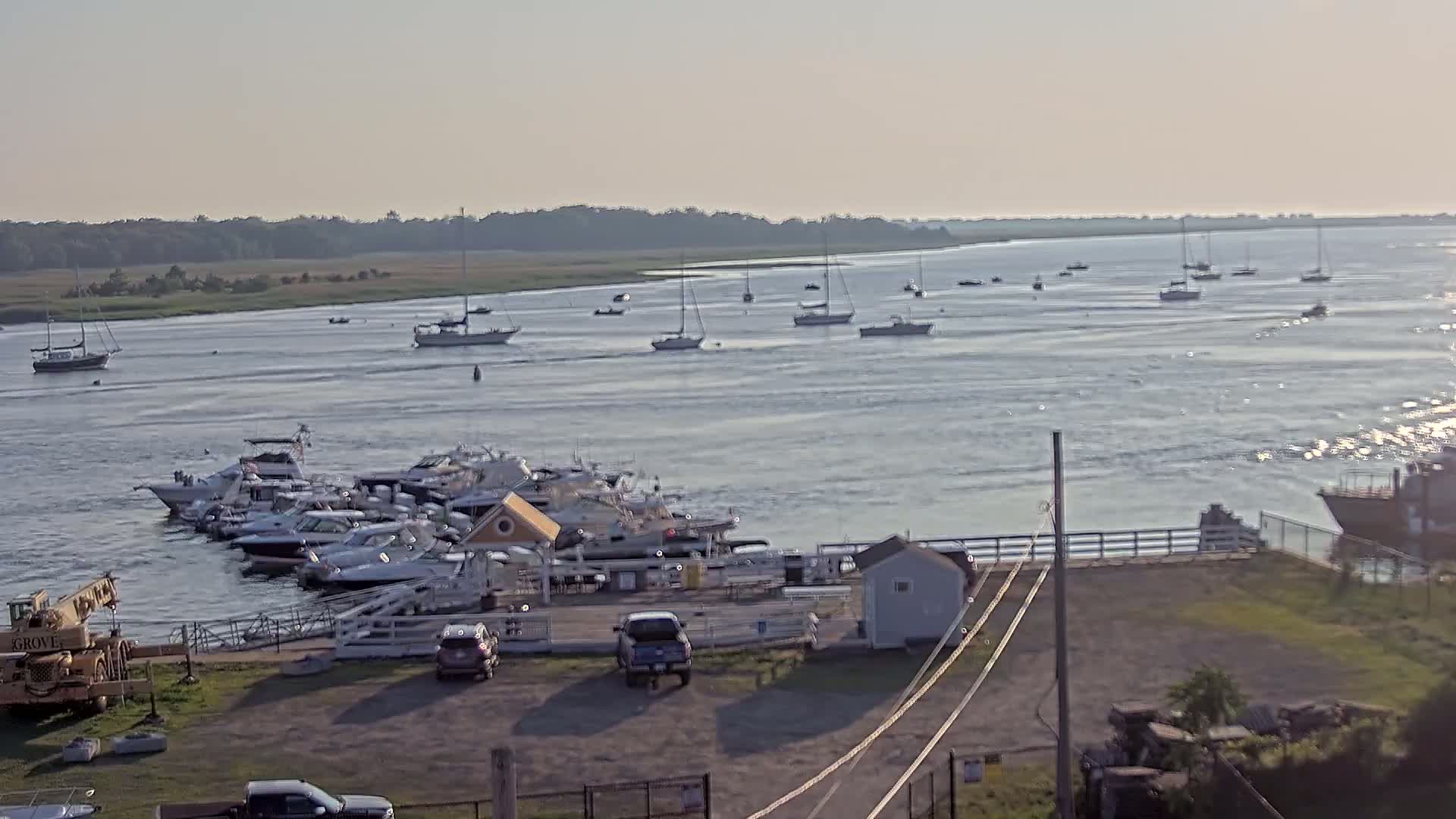A calm, sunny day reveals numerous boats docked at a marina, with additional boats sailing on a wide waterway bordered by greenery.
