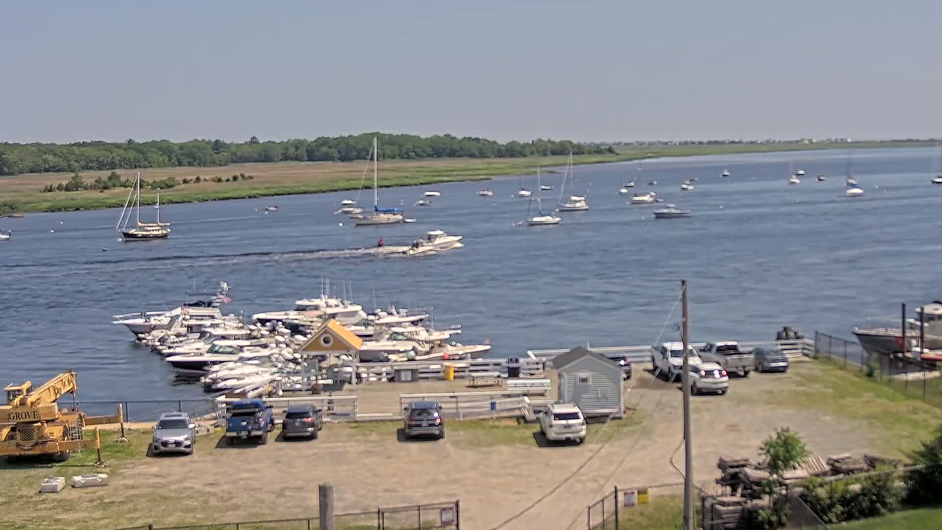 A marina with numerous boats docked, along with several cars parked in a lot, is situated on a calm, sunny day next to a wide body of water with additional boats scattered across its surface.