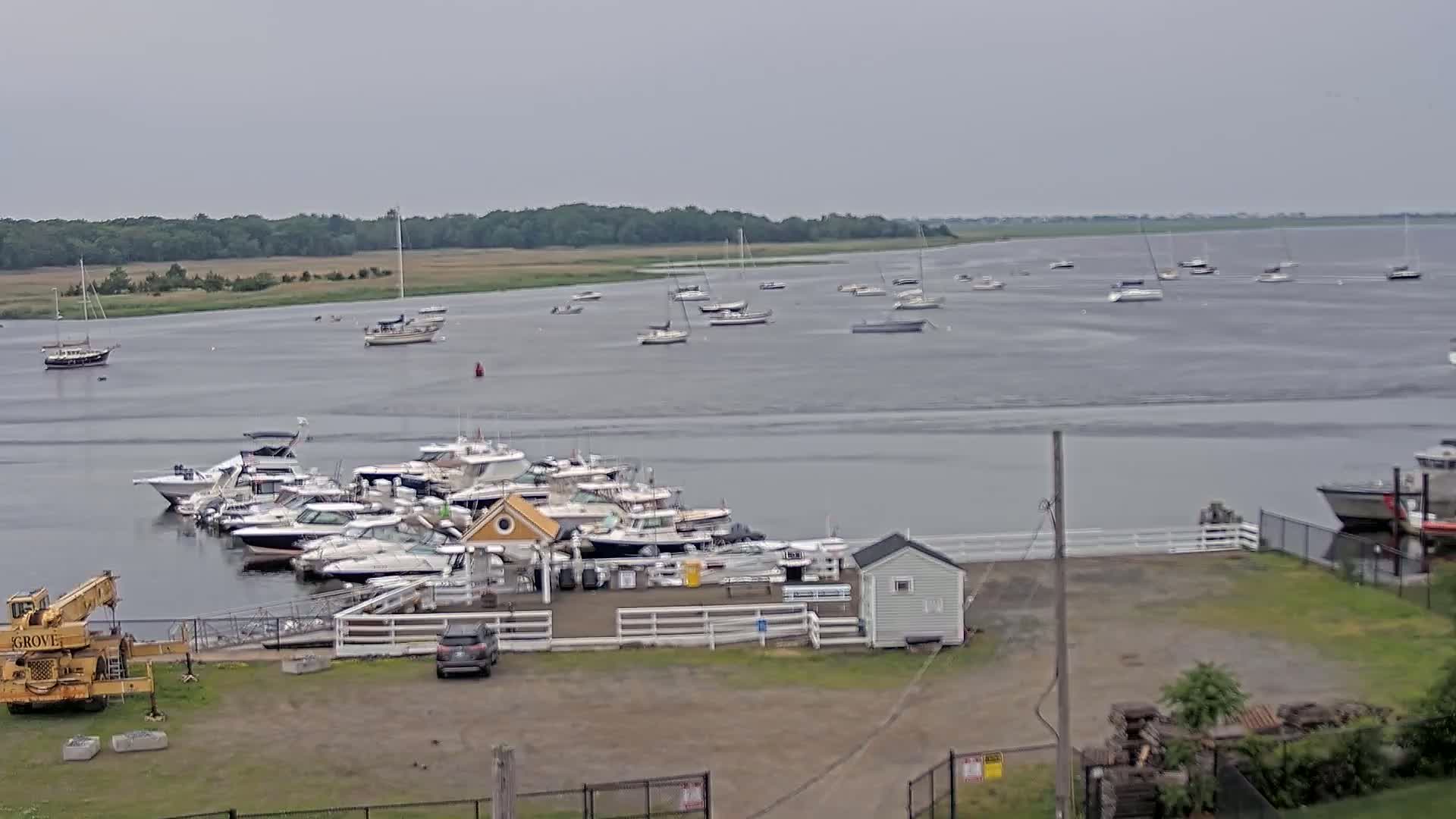 A marina with numerous boats docked, situated on a calm, gray day next to a body of water where additional boats are anchored.