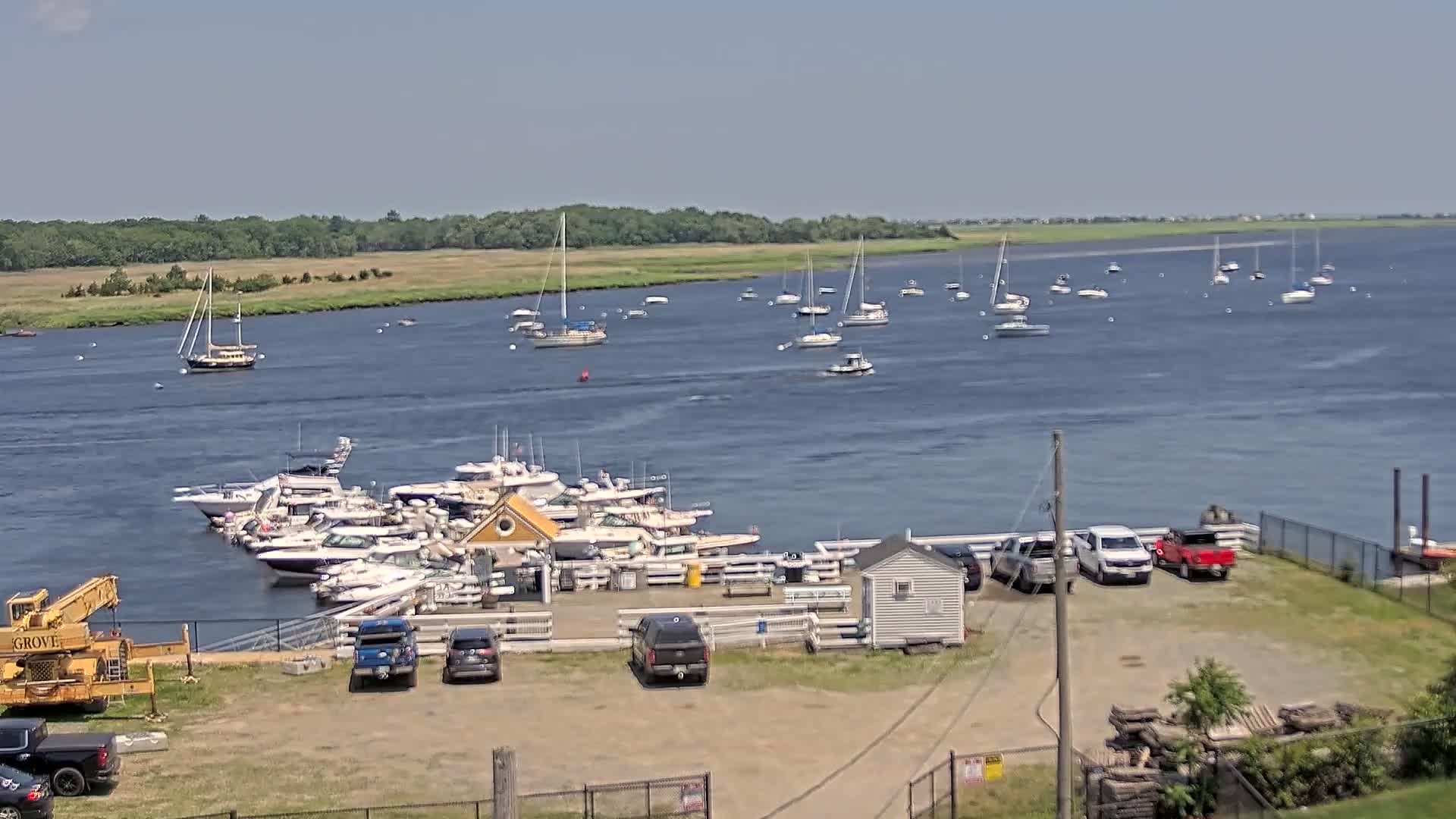 A marina filled with boats is situated on the shore of a calm body of water under a clear, sunny sky, with additional boats visible on the water in the distance.