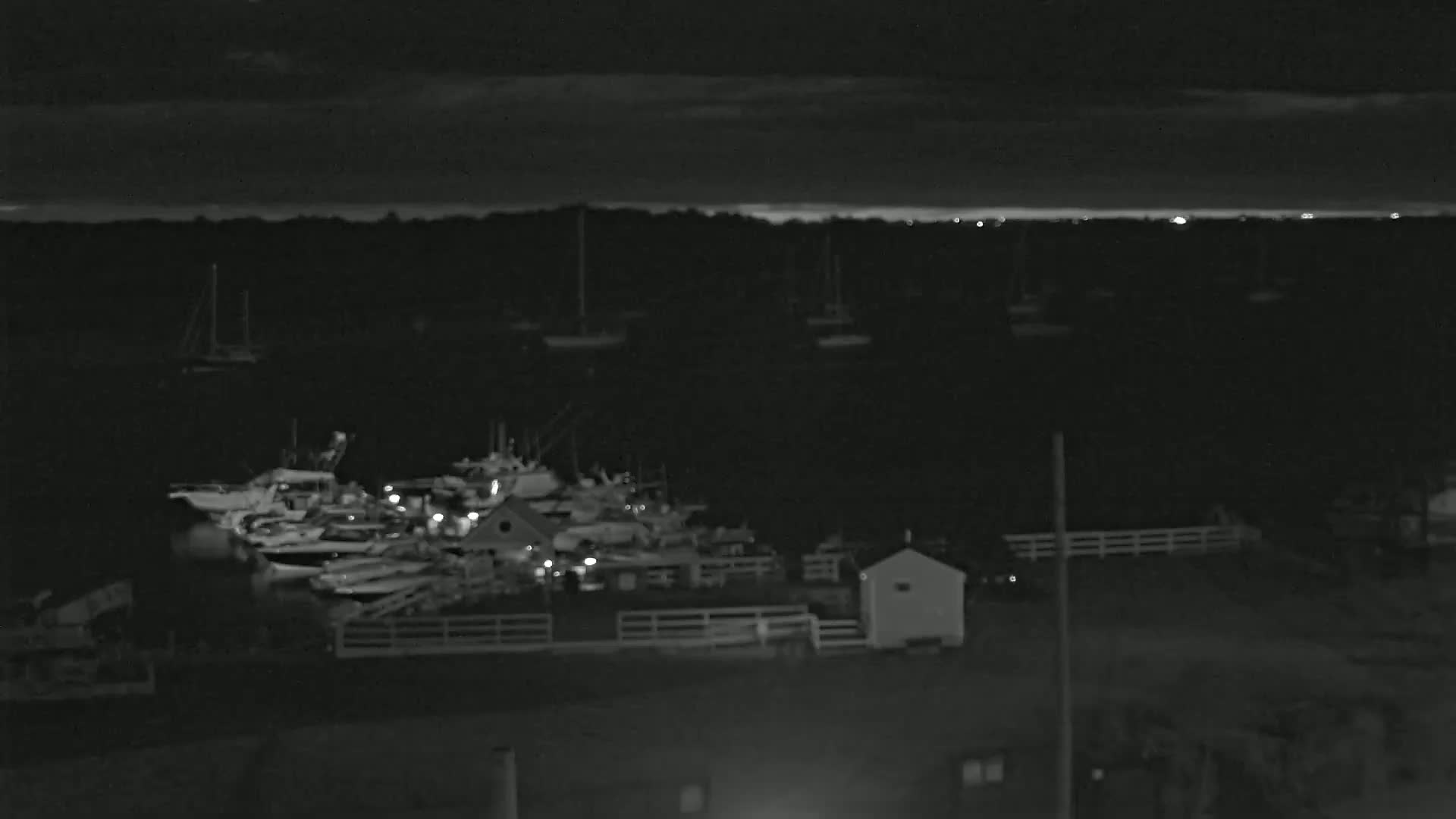 A nighttime view of a harbor with numerous boats docked, some lights visible, under a partly cloudy sky.