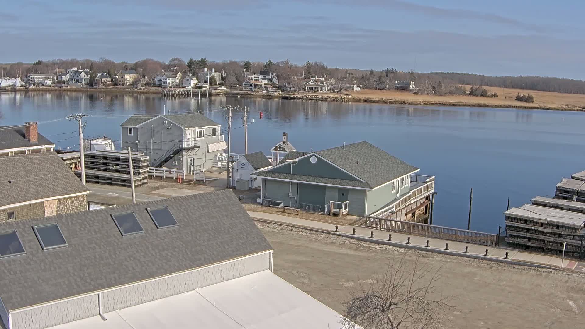 A waterfront view of a small town on a calm, sunny day, showing several houses, a few buildings near the water's edge, and stacks of what appear to be lobster traps.