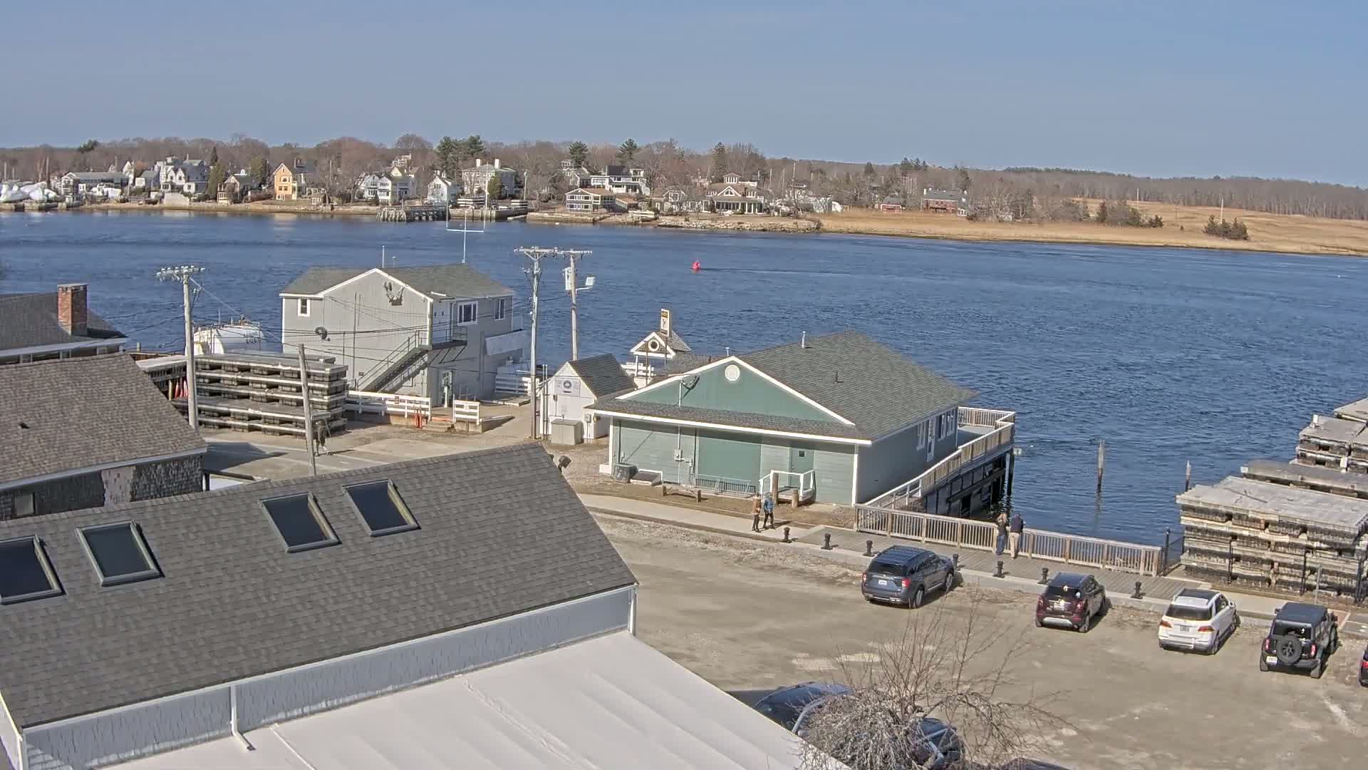 A waterfront scene on a sunny day shows several buildings, cars in a parking lot, and a body of water with houses and trees visible in the distance.