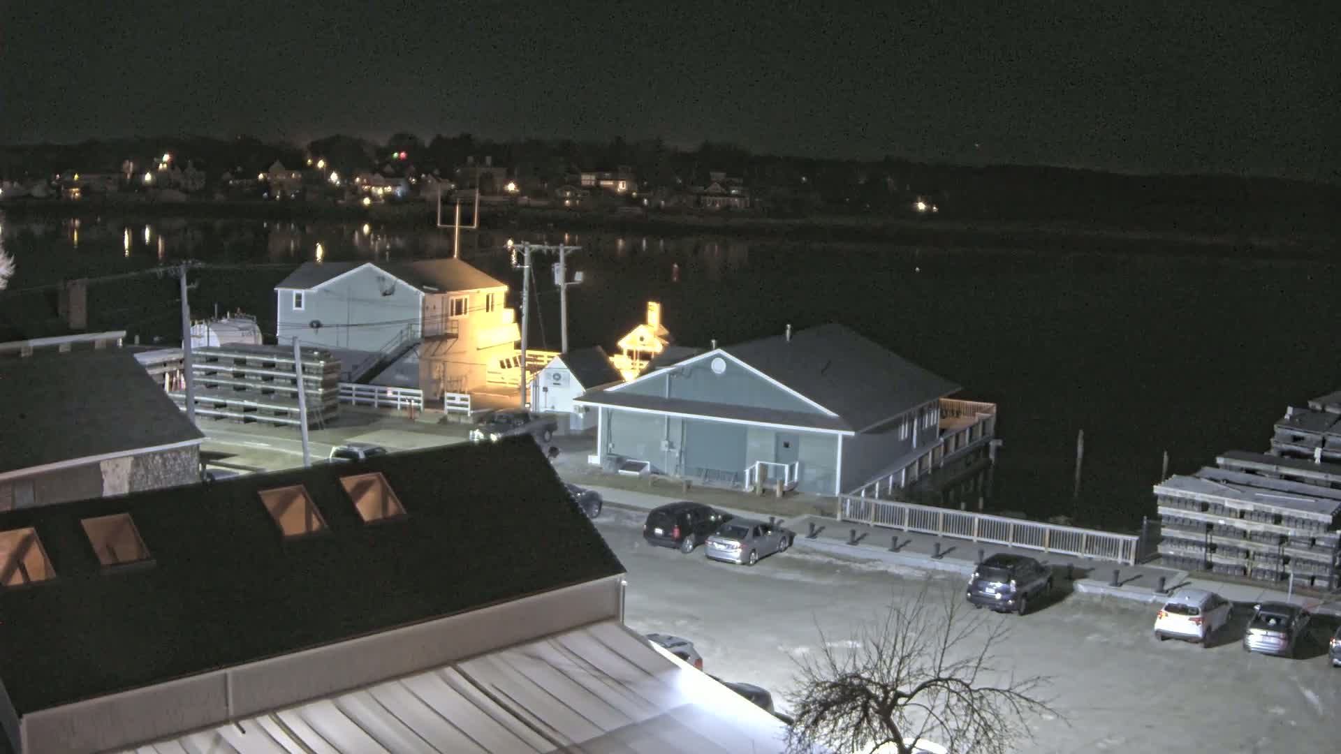 A nighttime view of a waterfront town shows several buildings, parked cars, and a calm body of water reflecting the lights of the buildings.
