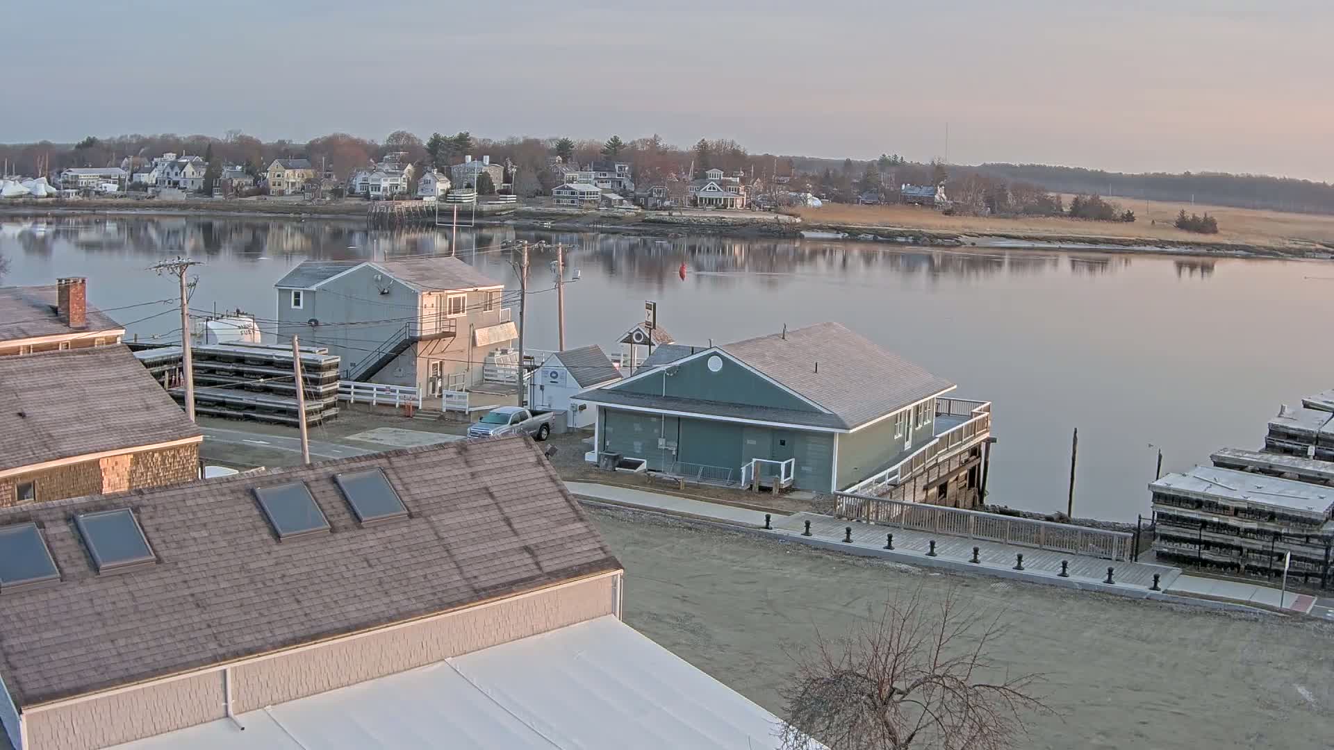 A waterfront view of a calm river reflecting a pale sunrise sky, showing several buildings, a parked pickup truck, and stacked floating docks.