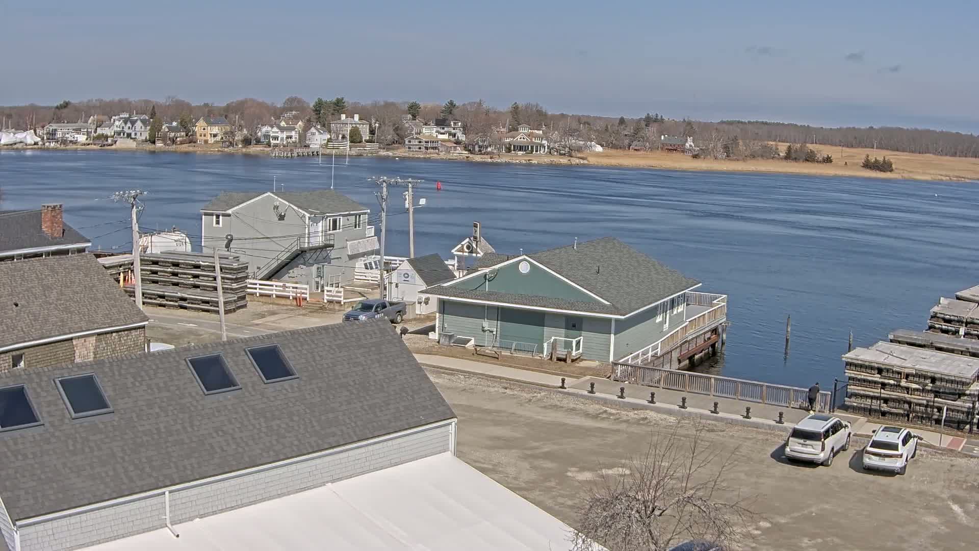 A waterfront view on a sunny day shows several buildings near a body of water, with cars parked in a lot and stacks of what appear to be wooden pallets or crates nearby.