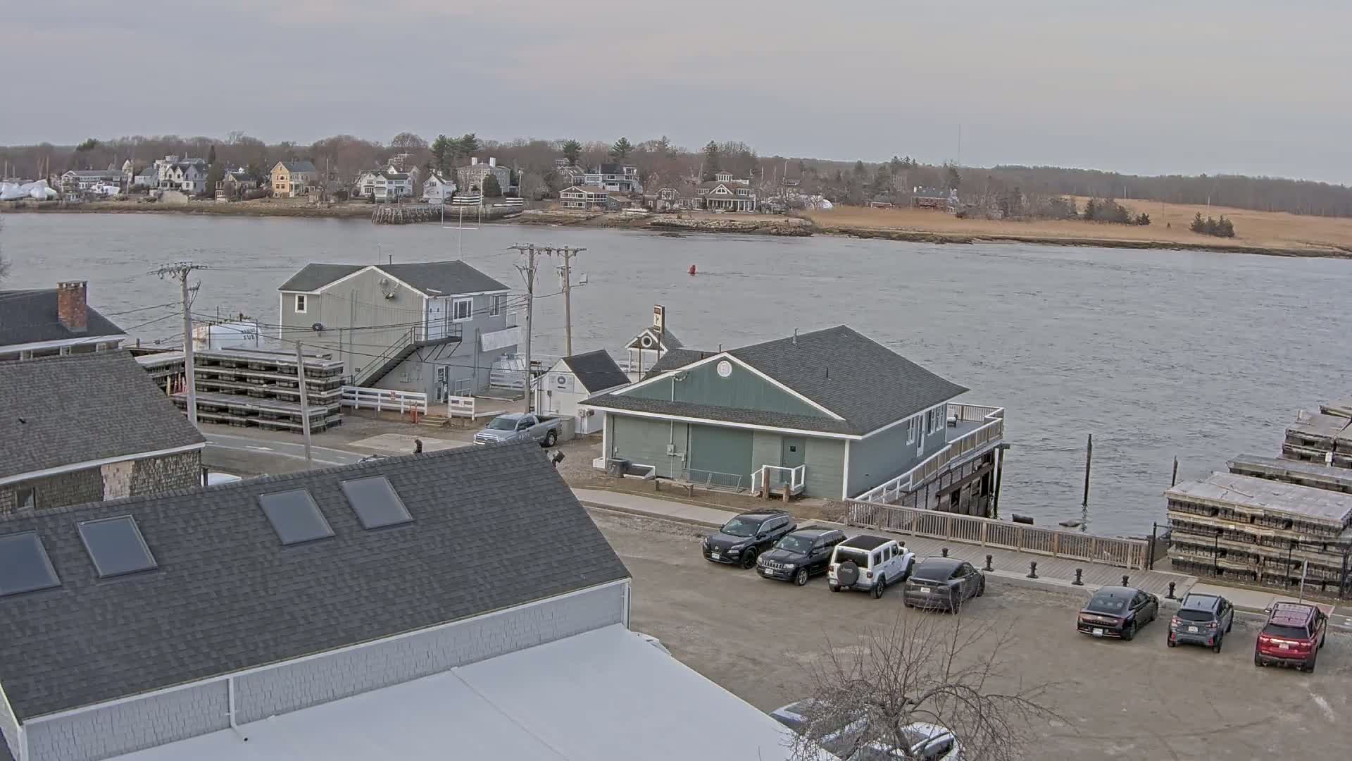 A waterfront scene on an overcast day shows several buildings, a parking lot with cars, and stacks of what appear to be lobster traps, with a calm river and a distant shoreline in the background.