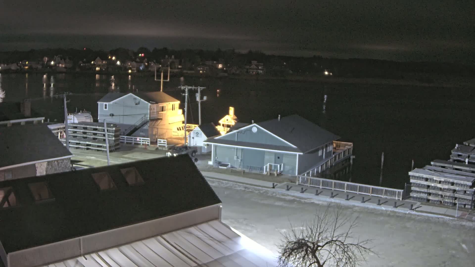 A nighttime view of a harbor, showing several buildings along a waterfront road, with stacks of what appear to be lobster traps, under a dark, clear sky.
