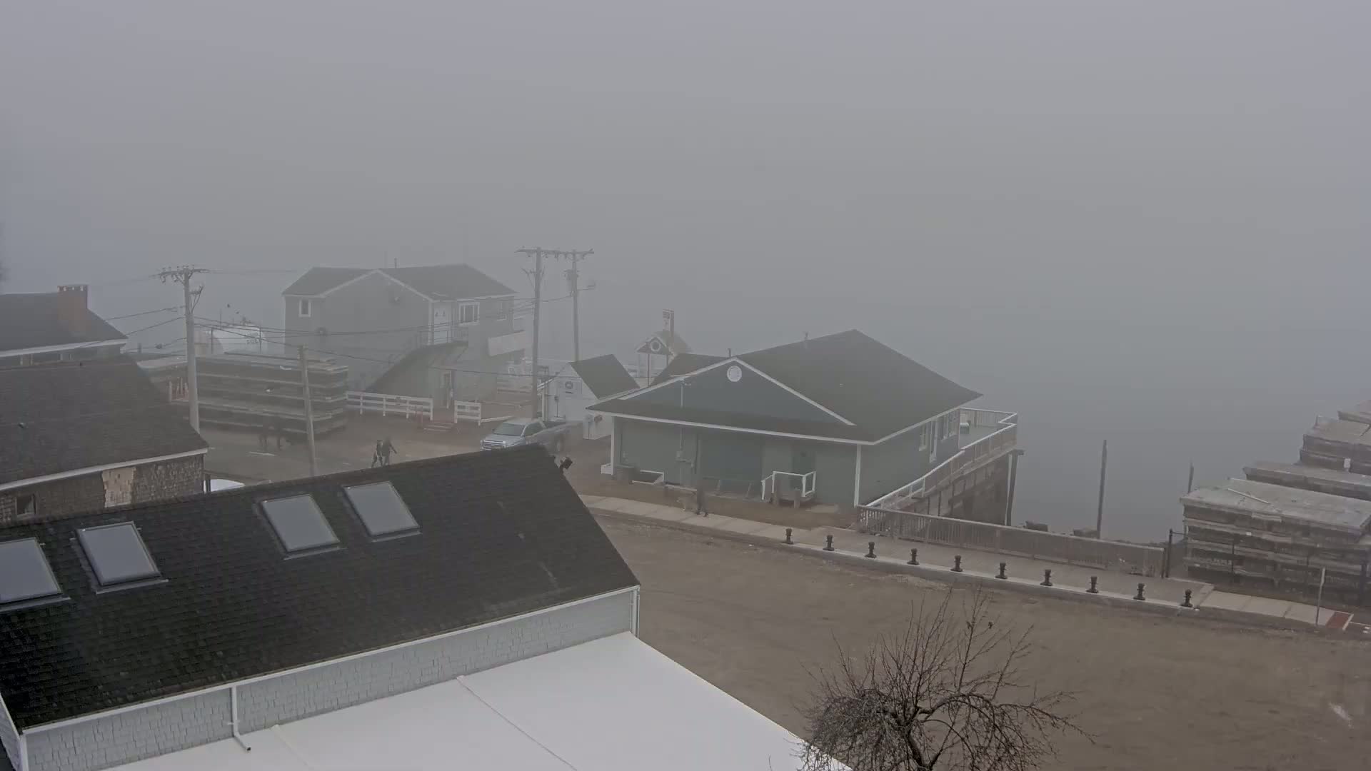 A foggy waterfront scene shows several houses and a dock with a few people walking around.