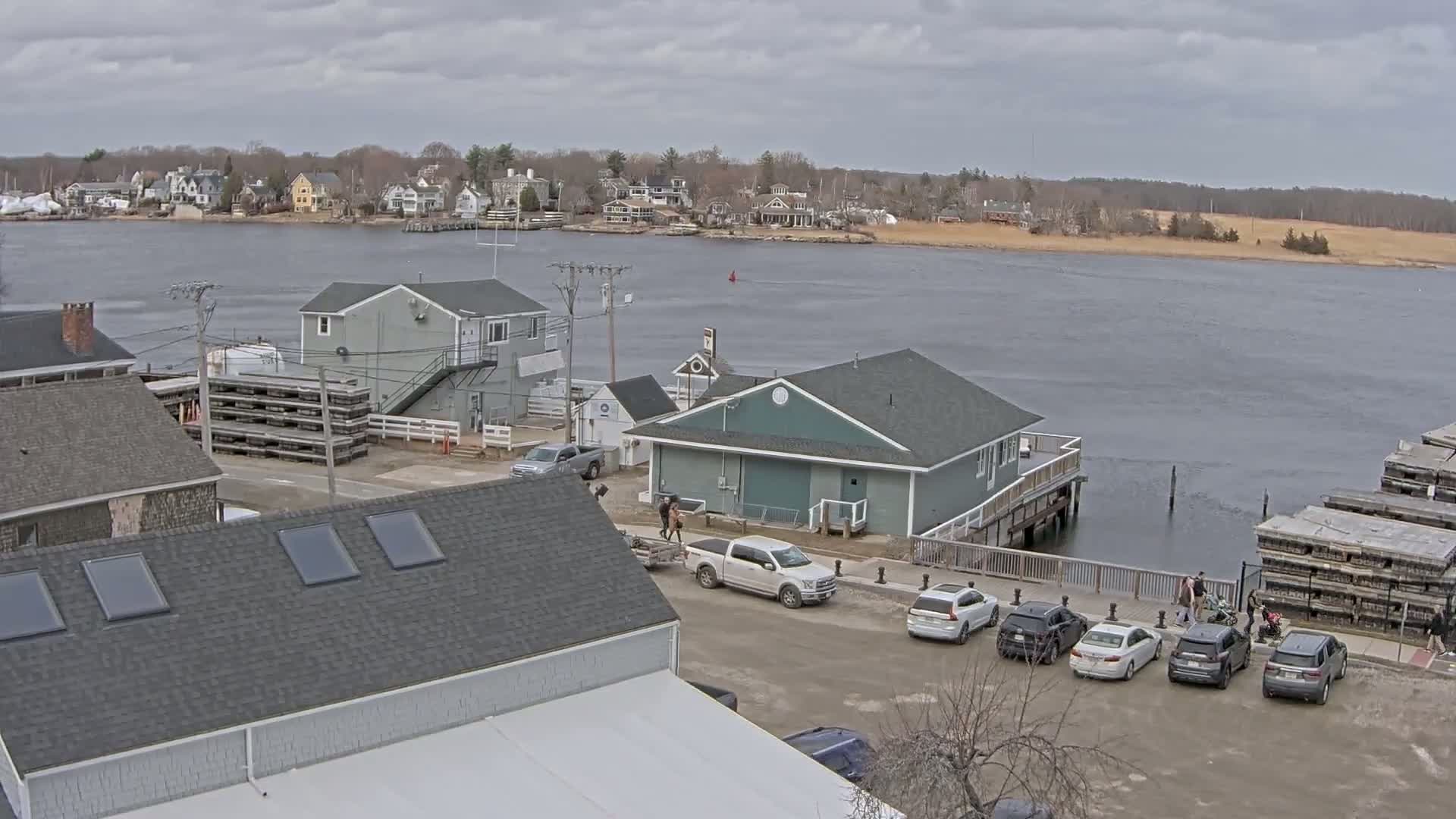 A waterfront scene on an overcast day shows several buildings, parked cars, and stacks of what appear to be oyster cages near a body of water with houses visible across it.