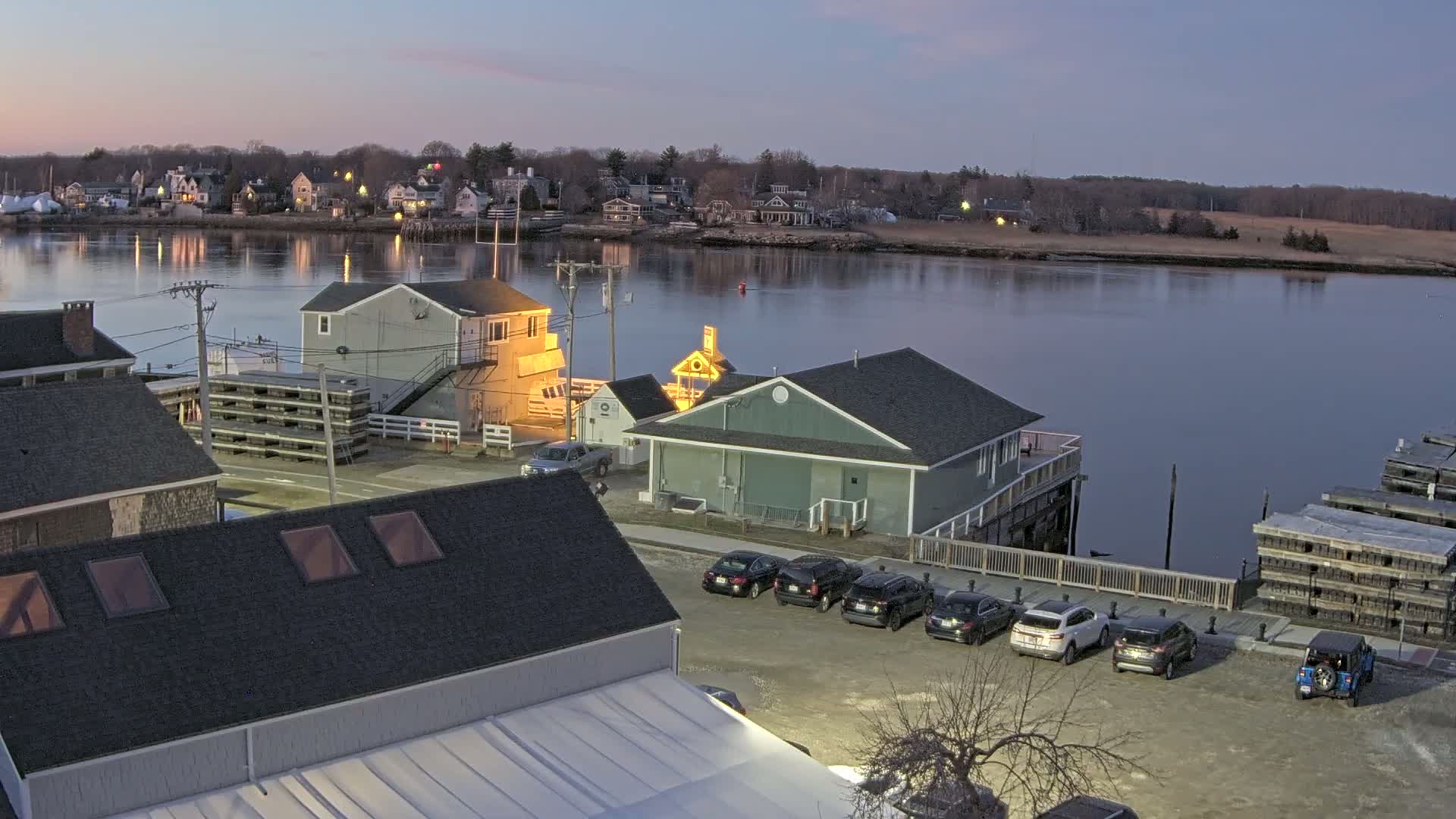 A waterfront town at dusk, with calm water reflecting the twilight sky, shows buildings, parked cars, and lobster traps.