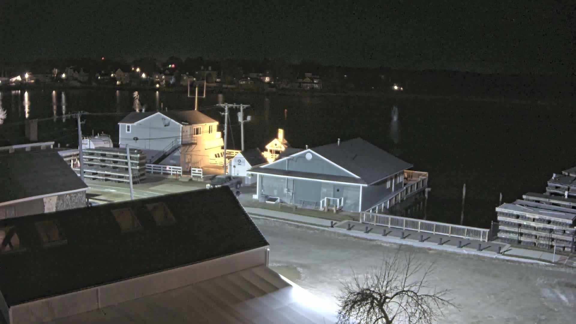 A nighttime view of a waterfront area with several buildings, a body of calm water, and stacks of what appear to be lobster traps.