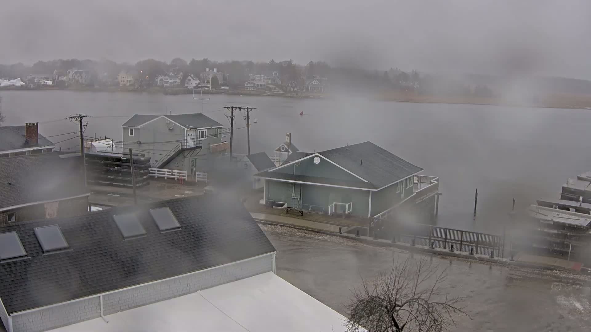 A foggy, rainy scene shows several buildings near a body of water that has overflowed onto a street, partially submerging a small dock and a portion of the roadway.