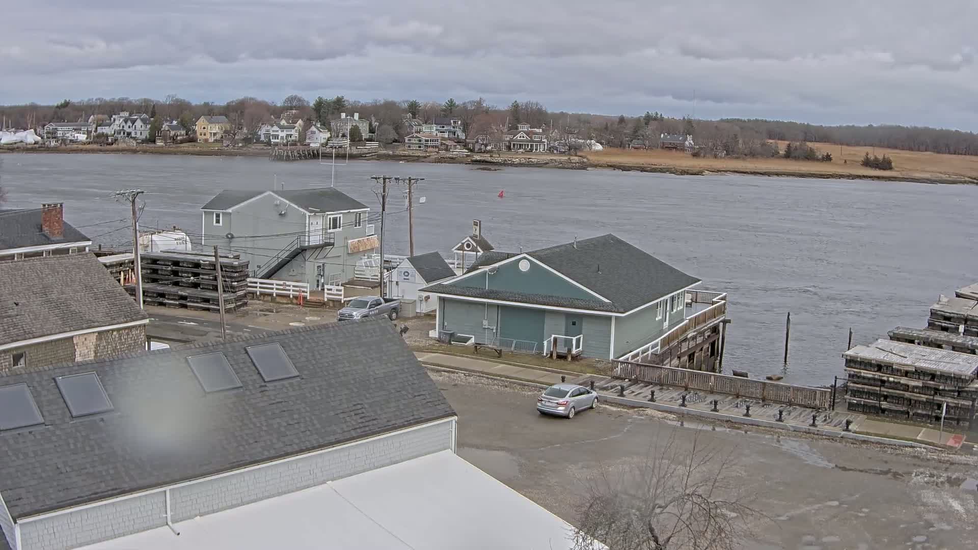 A waterfront scene on an overcast day shows a gray building near a river with houses and trees in the background, a few cars in a parking lot, and stacks of wooden structures near the water.