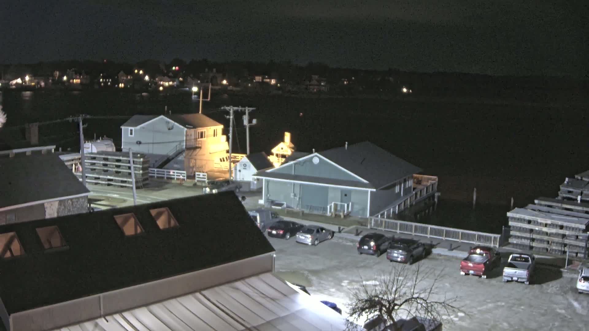 A nighttime aerial view of a waterfront area shows several buildings, a parking lot with cars, and stacks of wooden planks near the water's edge.
