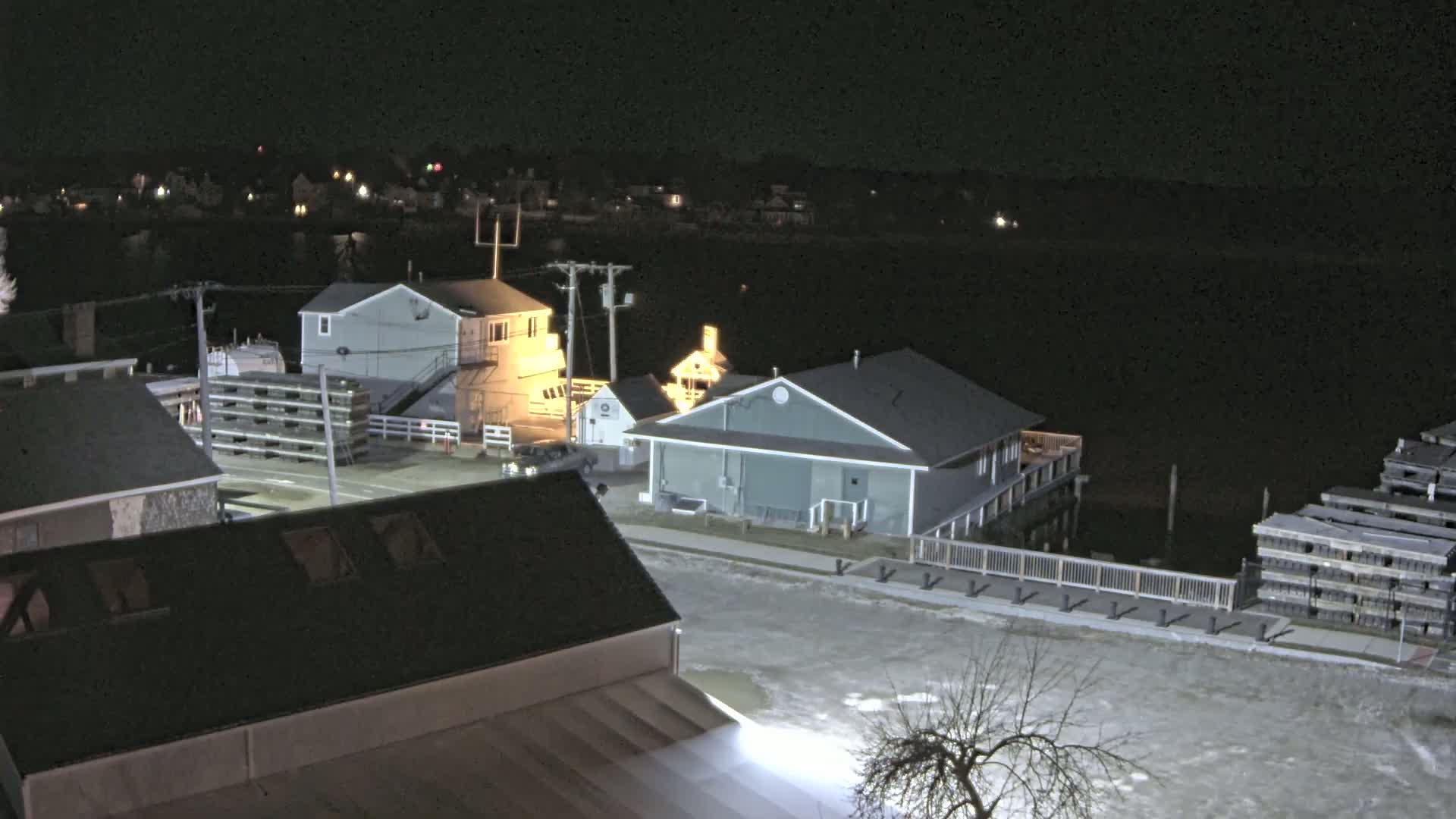 A nighttime view of a waterfront area with several buildings, stacks of what appear to be wooden pallets, and a partially frozen or snow-covered ground.
