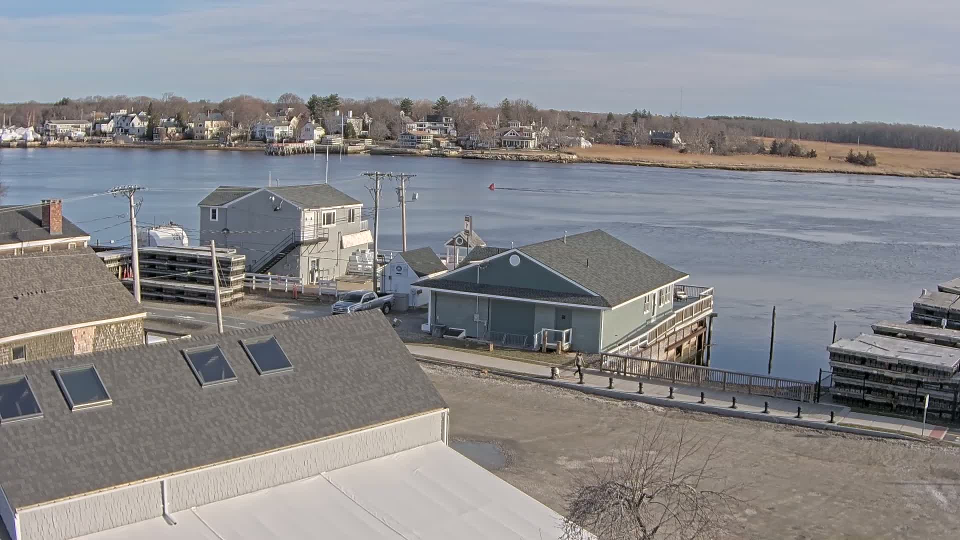A waterfront town scene on a sunny day shows several buildings near a calm river with some ice, with a residential area visible across the water.