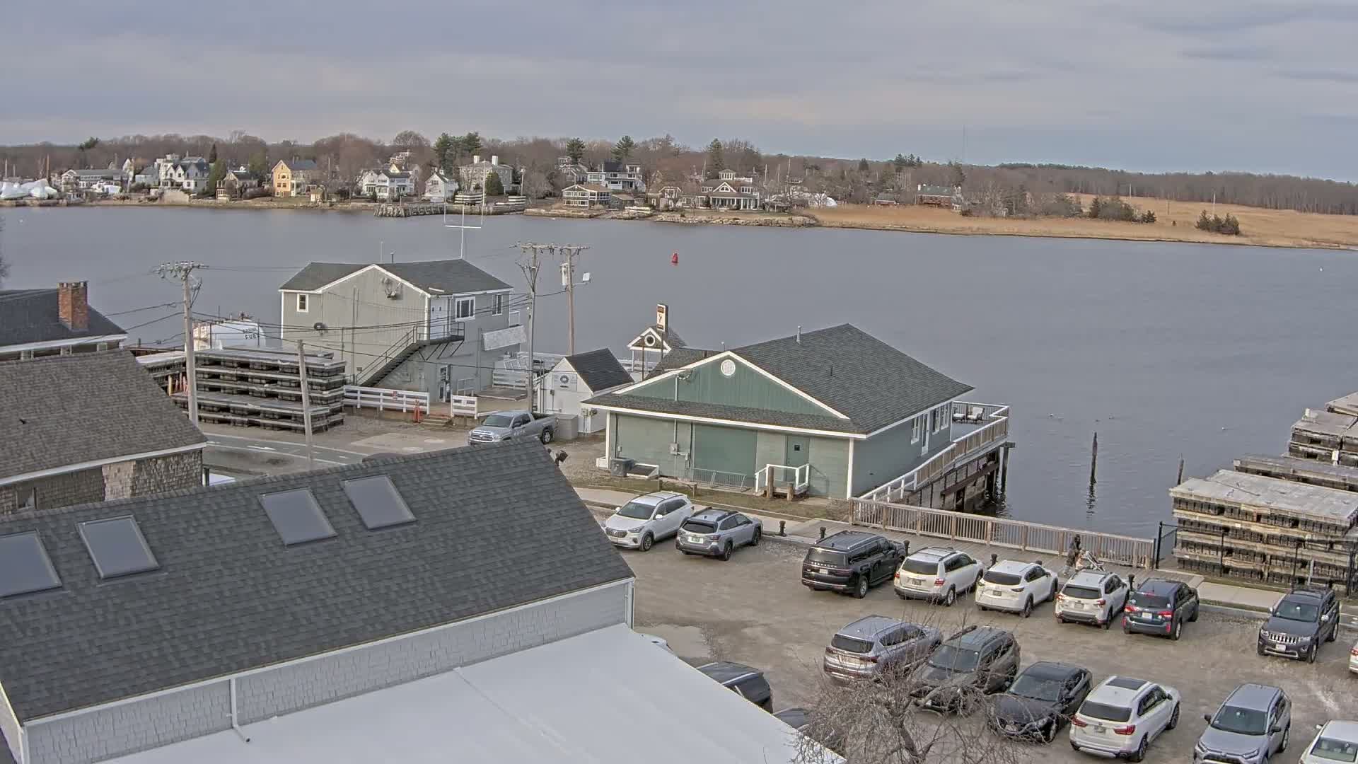 A waterfront scene on an overcast day shows several buildings, a parking lot full of cars, and stacks of what appear to be lobster traps near a calm body of water with a town visible in the distance.