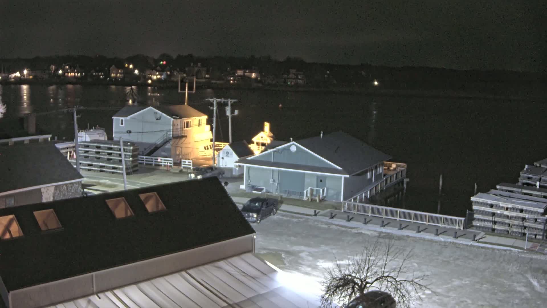 A nighttime aerial view of a waterfront town shows several buildings, a dark body of water, and snow or ice on the ground.