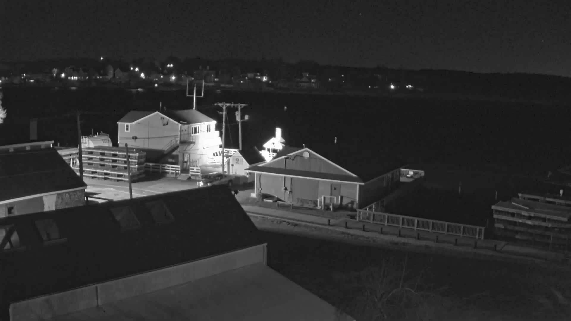 A nighttime, black-and-white view of a waterfront area with several buildings, a body of water, and stacked wooden pallets.