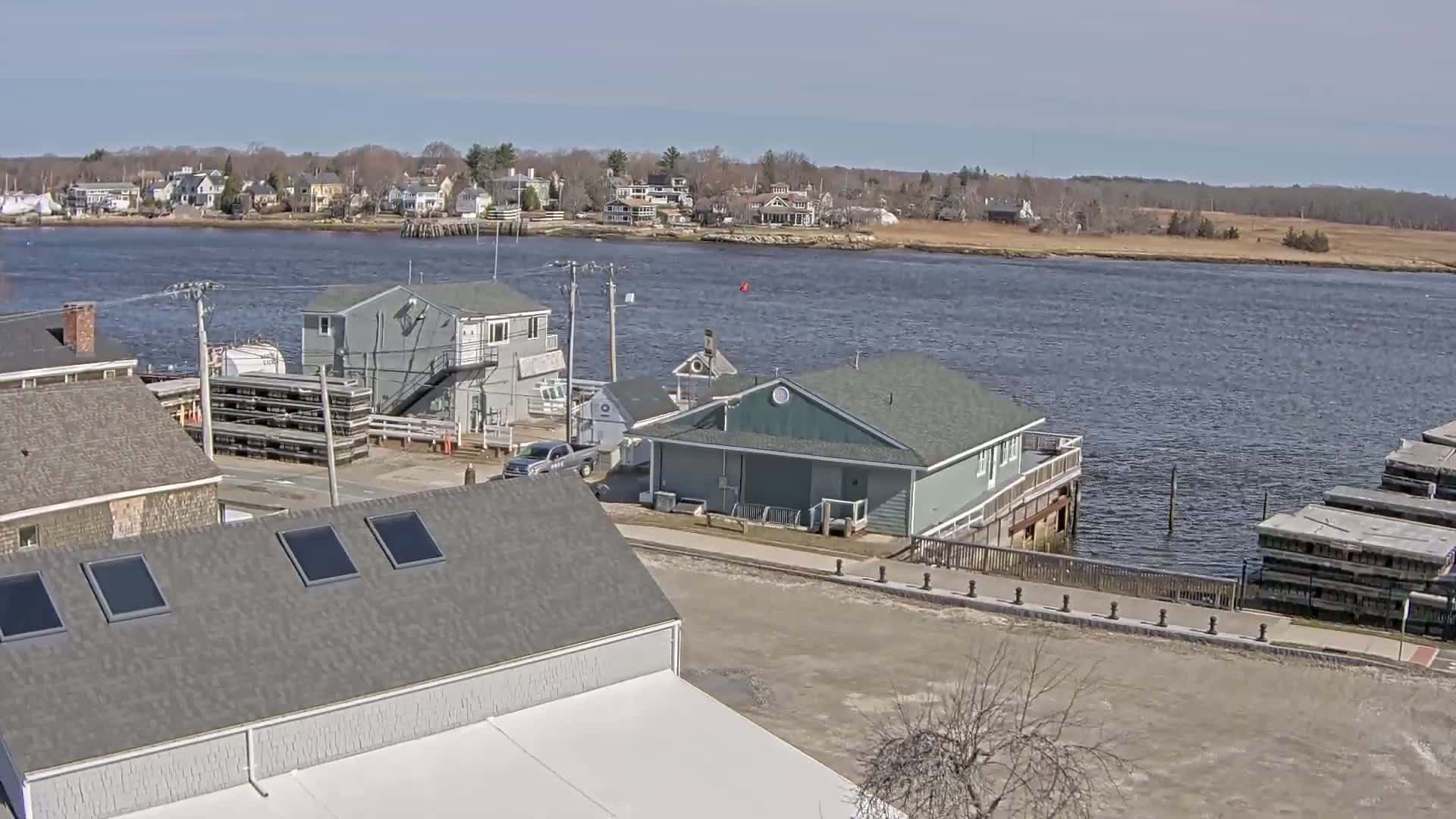 A waterfront view on a sunny day shows several buildings near a calm river with a town in the background.