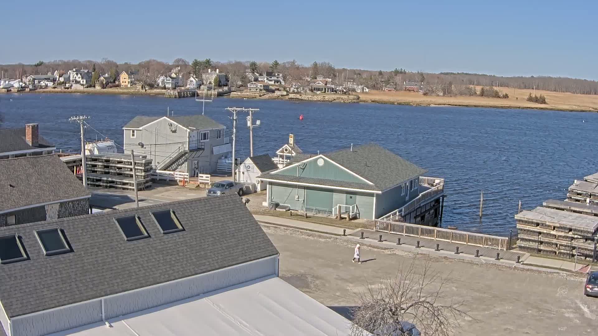 A waterfront village scene on a sunny day shows several buildings near a calm body of water with a far shoreline of trees and houses.