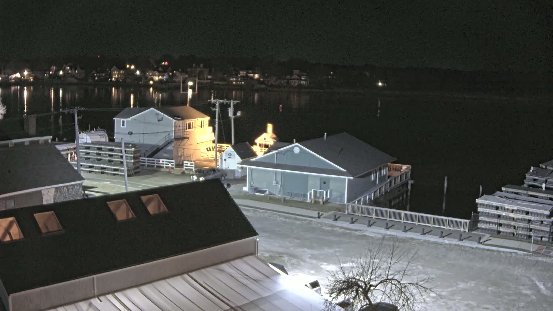 A nighttime, snow-covered waterfront scene shows several buildings near a body of water, with lights illuminating the area.