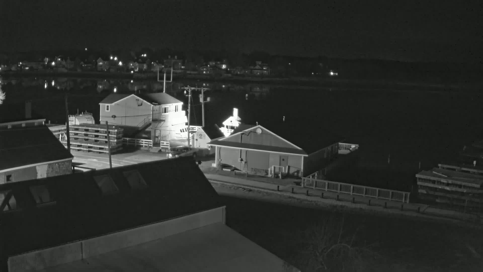 A nighttime, black-and-white view of a waterfront area with buildings, a dock, and a calm body of water reflecting the lights of a nearby town.