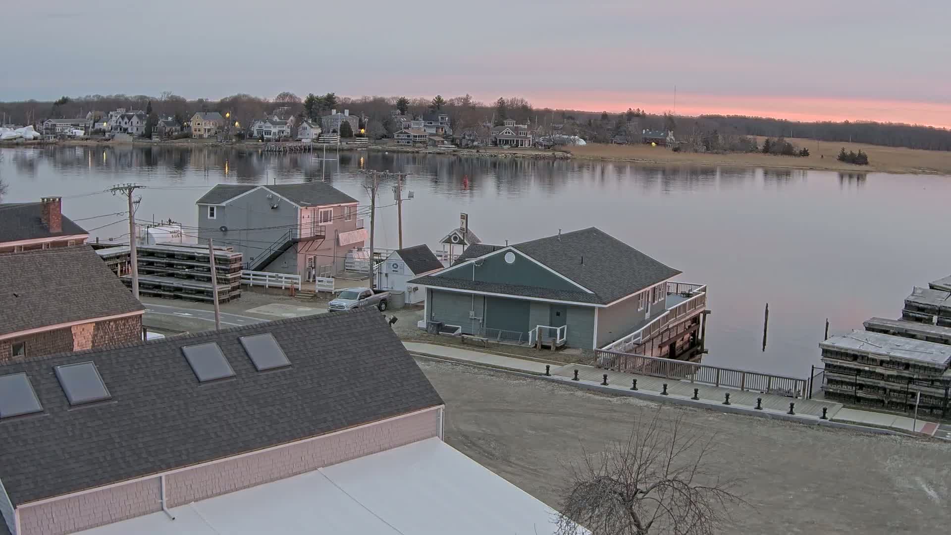 A waterfront village at dawn shows several buildings near a calm river with a pink sky.
