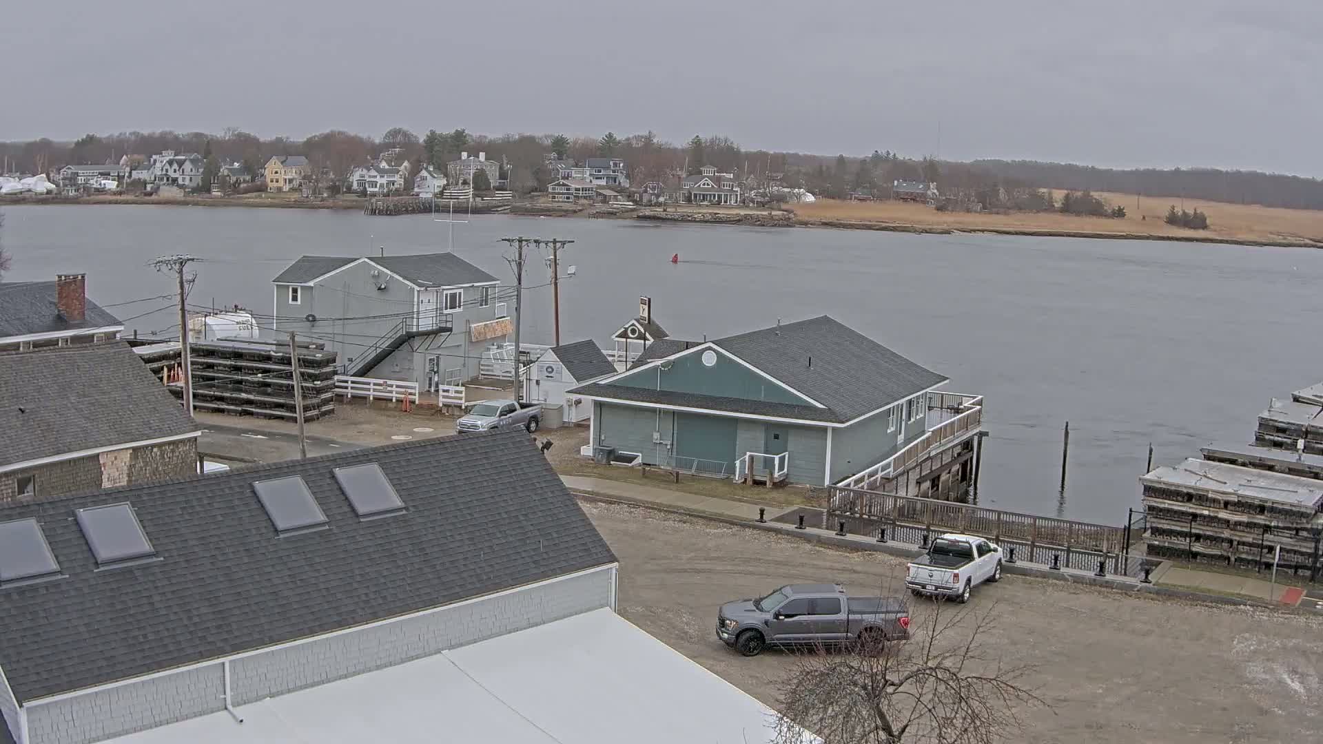 A waterfront scene on an overcast day shows several buildings, parked trucks, and a calm body of water with a distant shoreline of houses and trees.