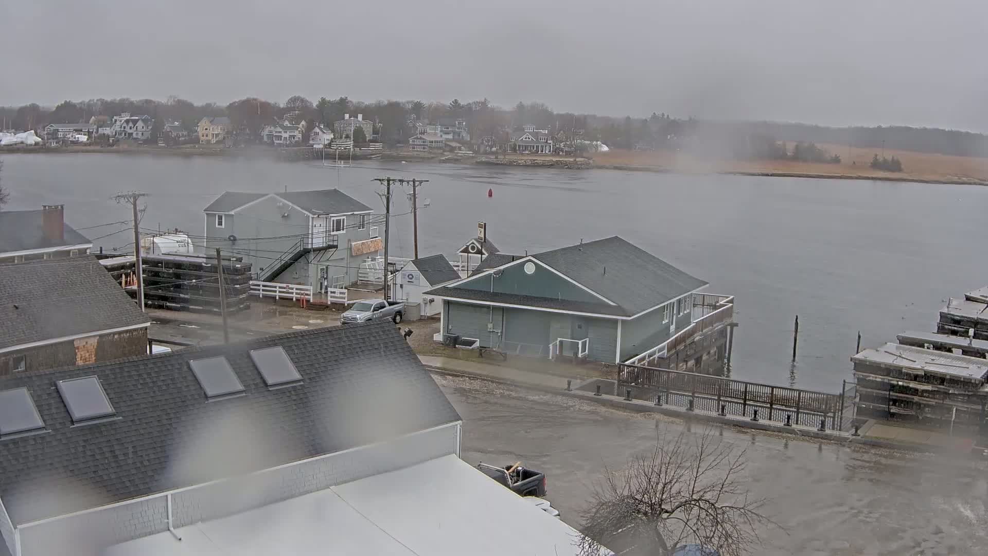 A waterfront scene on an overcast day shows several houses and buildings near a body of water, with some standing water in the street.