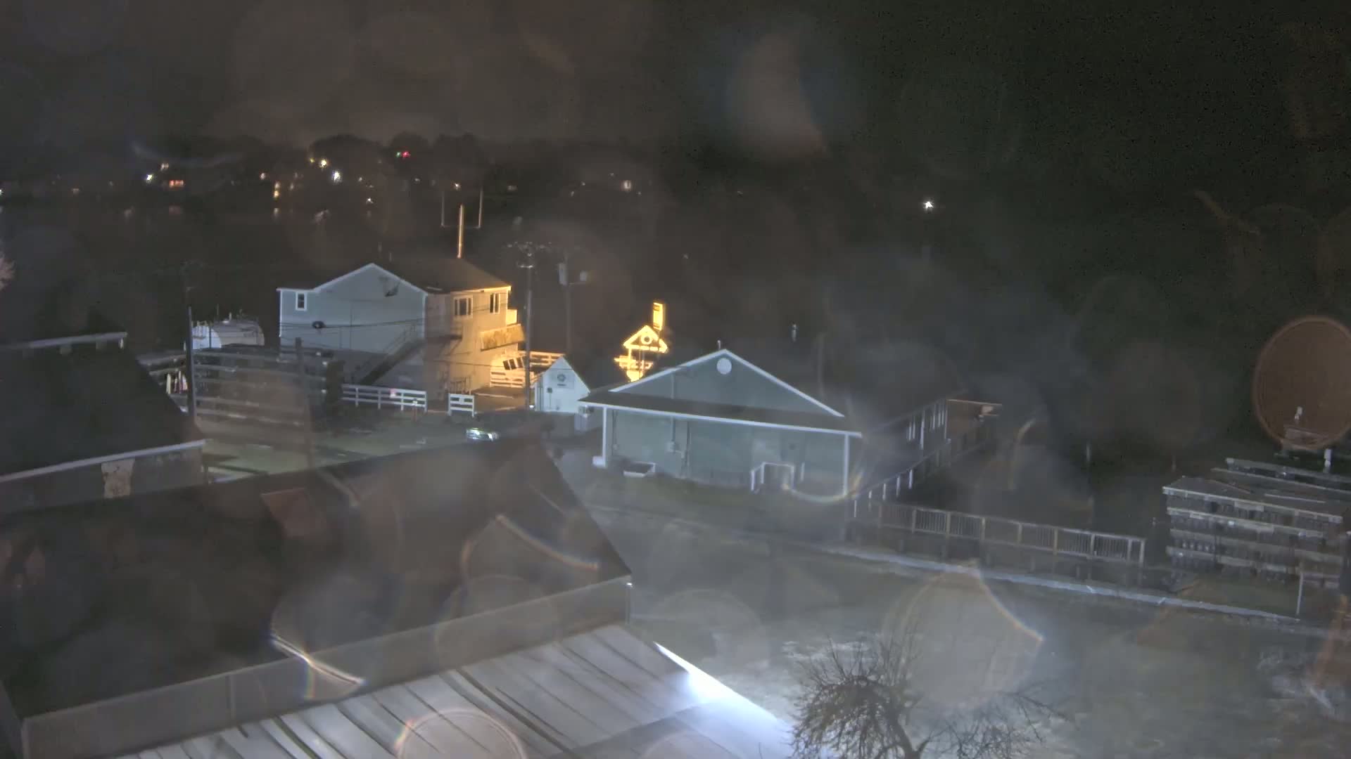 A nighttime view of several houses and buildings near a body of water, obscured by rain or mist.