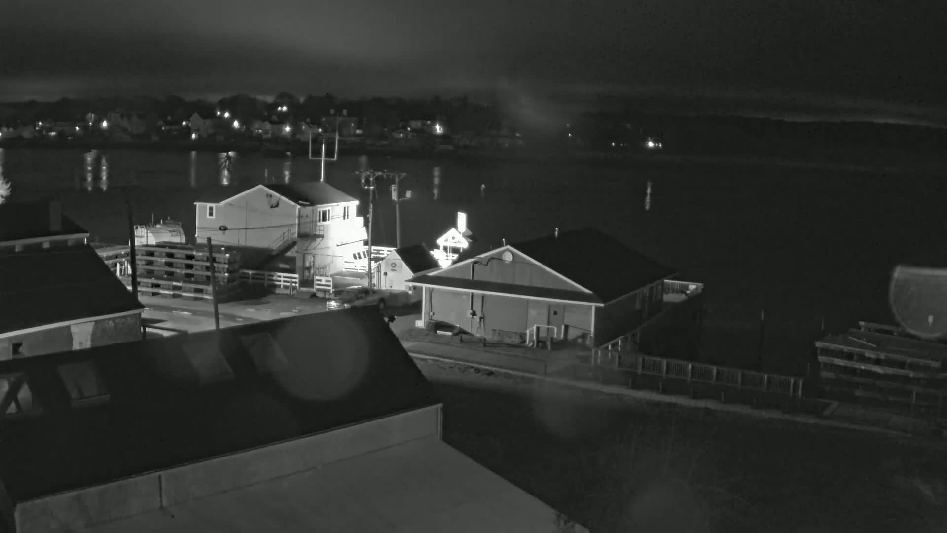 A nighttime, low-light view of several buildings near a calm body of water, with a town visible in the distance under an overcast sky.