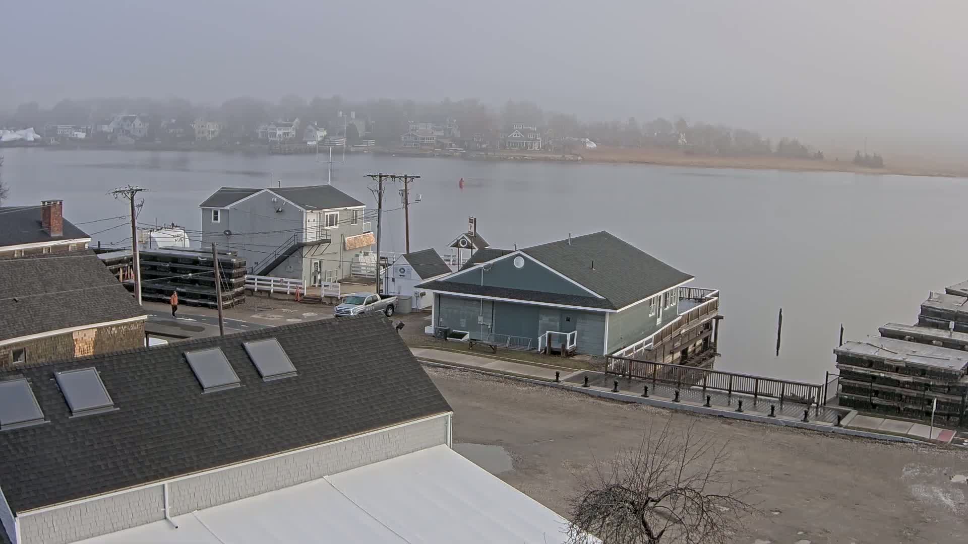 A foggy view of a waterfront town shows several buildings, a calm body of water, and a few vehicles parked on a street.