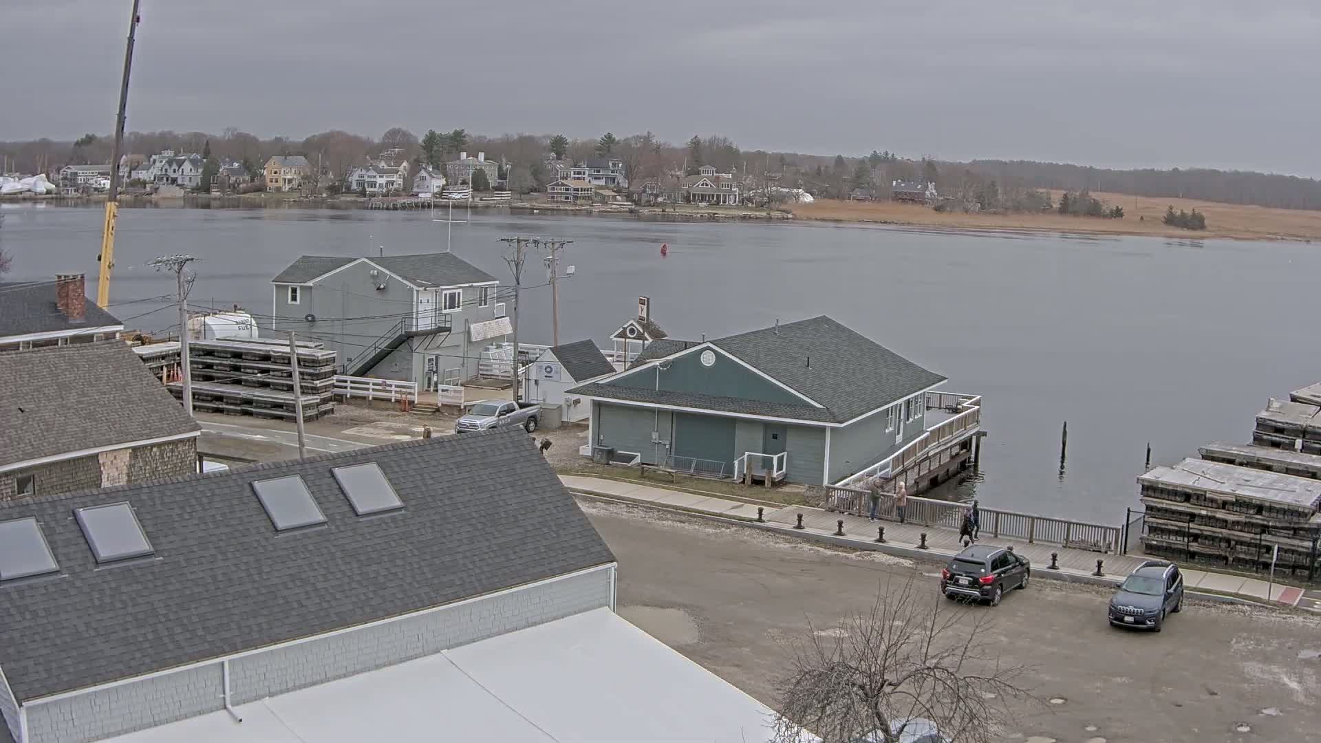 A waterfront scene on an overcast day shows several buildings, including a gray two-story house and a teal single-story building on a dock, with several cars parked nearby, and a body of water with houses across it.