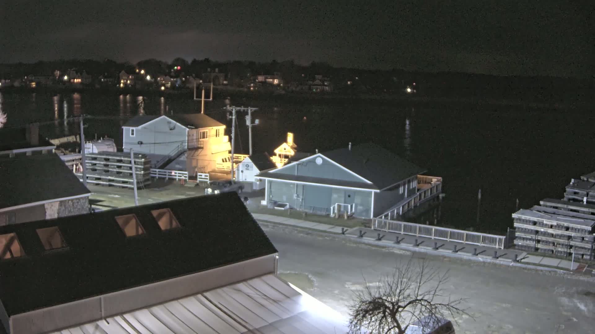 At night, a waterfront scene shows several buildings and a calm body of water reflecting lights from the opposite shore.