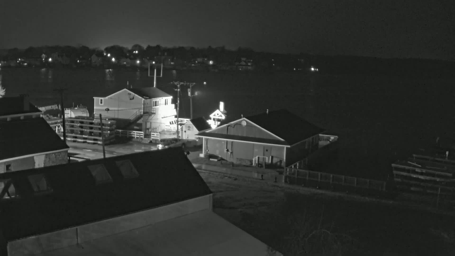 A nighttime, black and white aerial view shows several buildings near a body of water under dark, overcast skies.