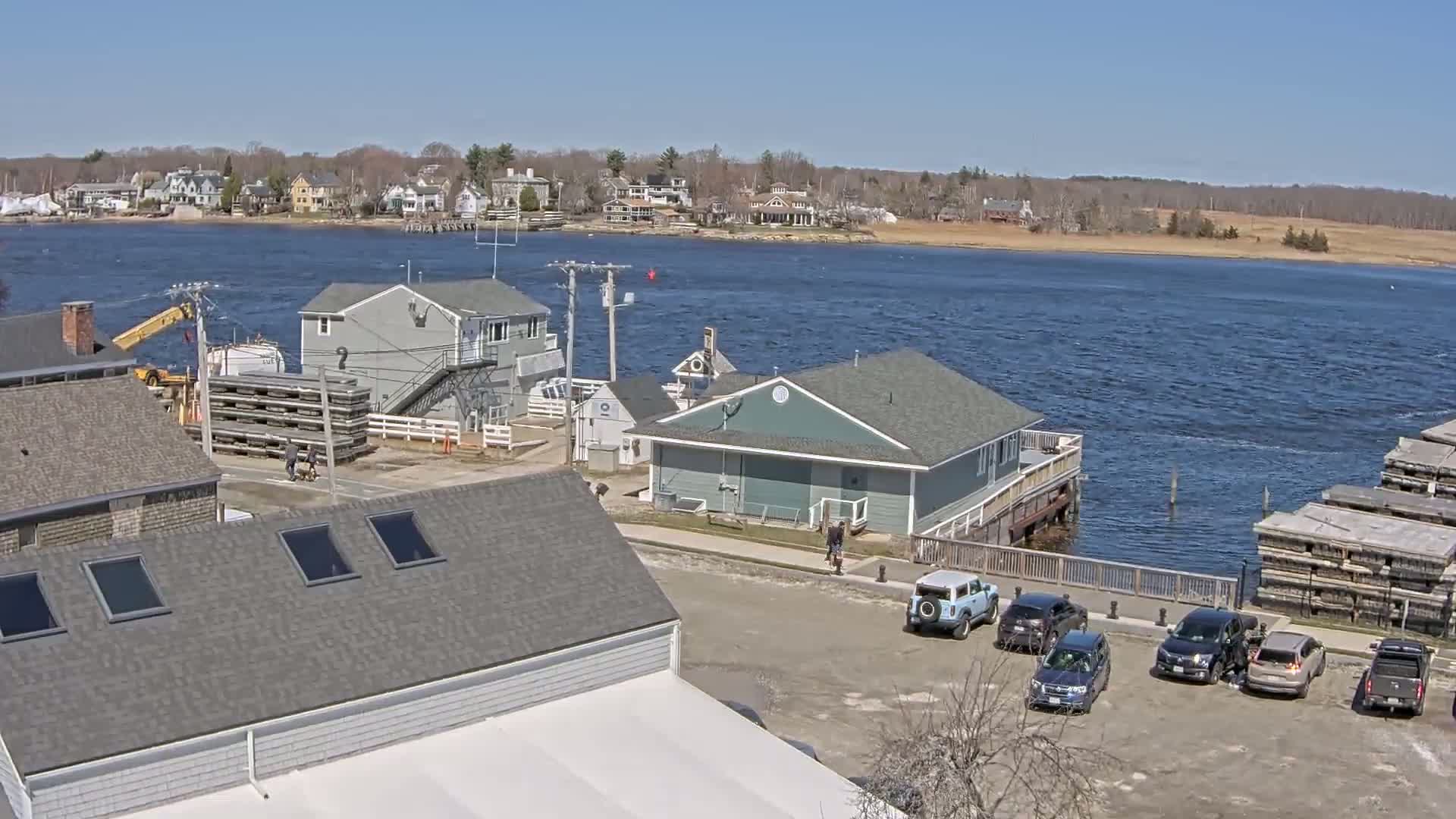 A waterfront scene on a sunny day shows a parking lot with several cars, several buildings near the water, and a body of water with small waves.