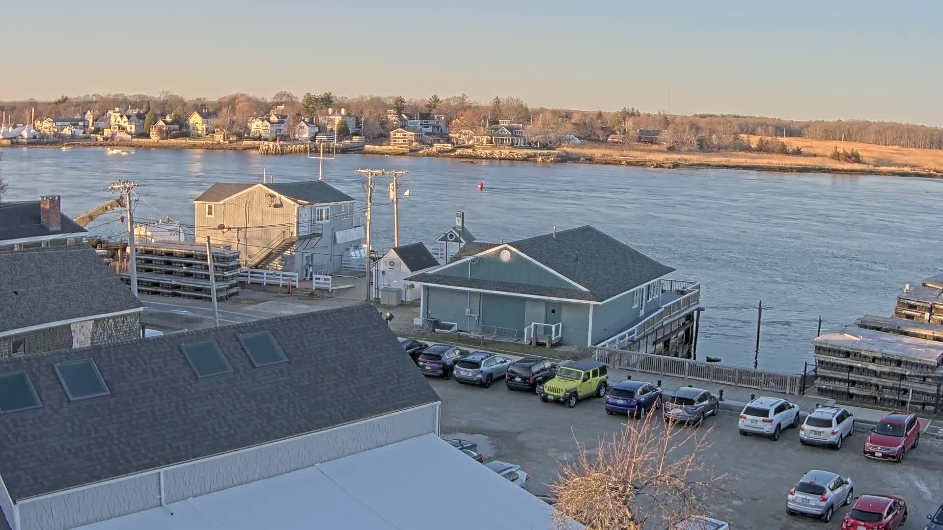 A waterfront scene on a calm, sunny day shows several buildings, parked cars, and a river with a town in the distance.