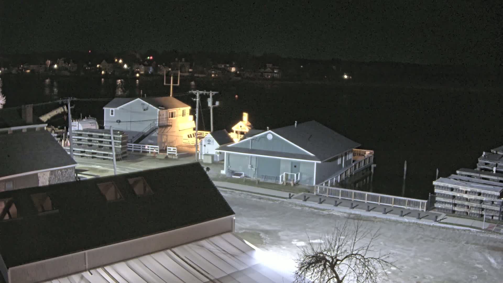 A nighttime view of a waterfront area with several buildings, a body of water, and stacks of what appear to be wooden structures, under clear, dark skies.