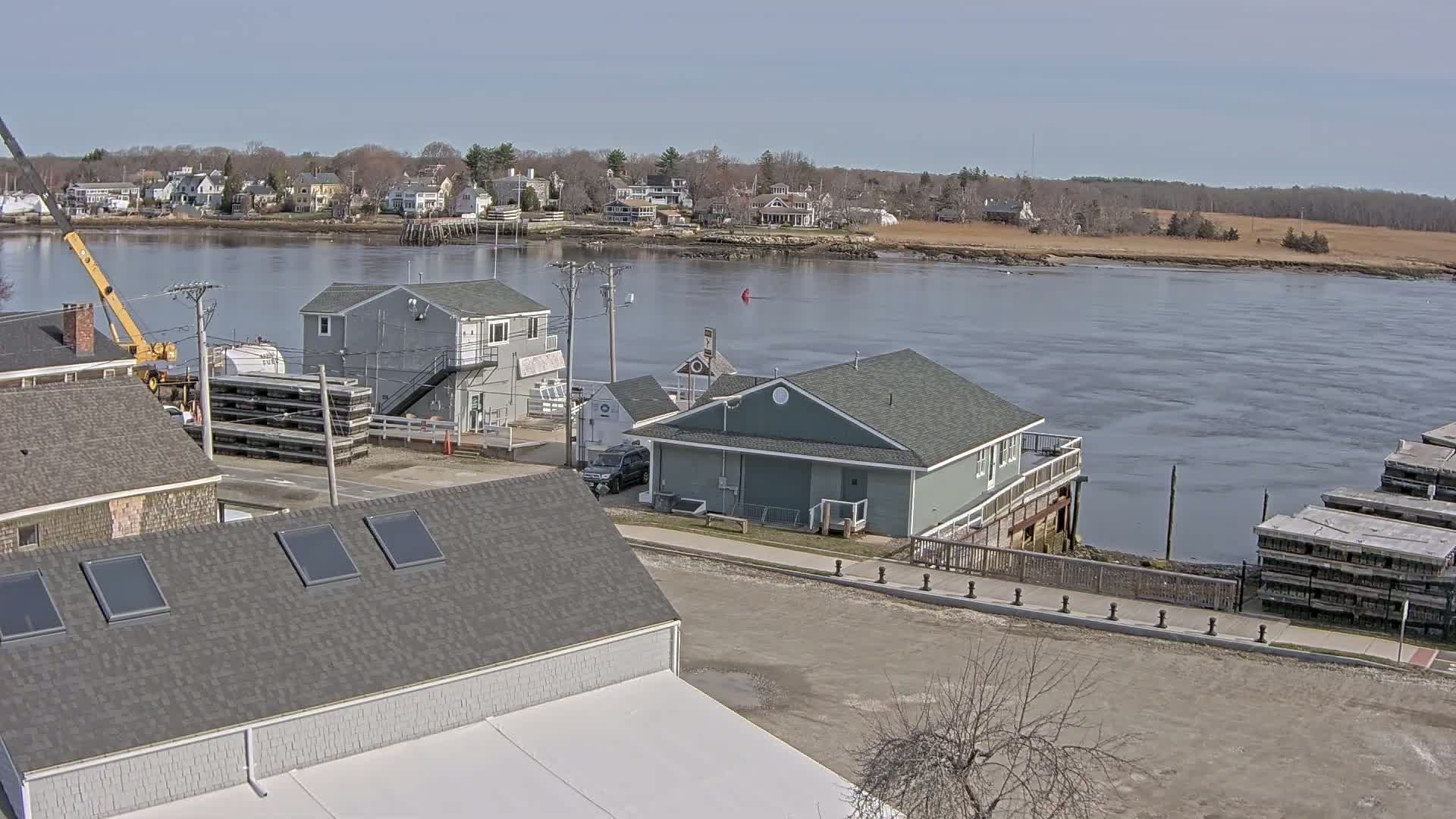 A waterfront view on a sunny day shows several buildings near a calm river with a town visible across the water.
