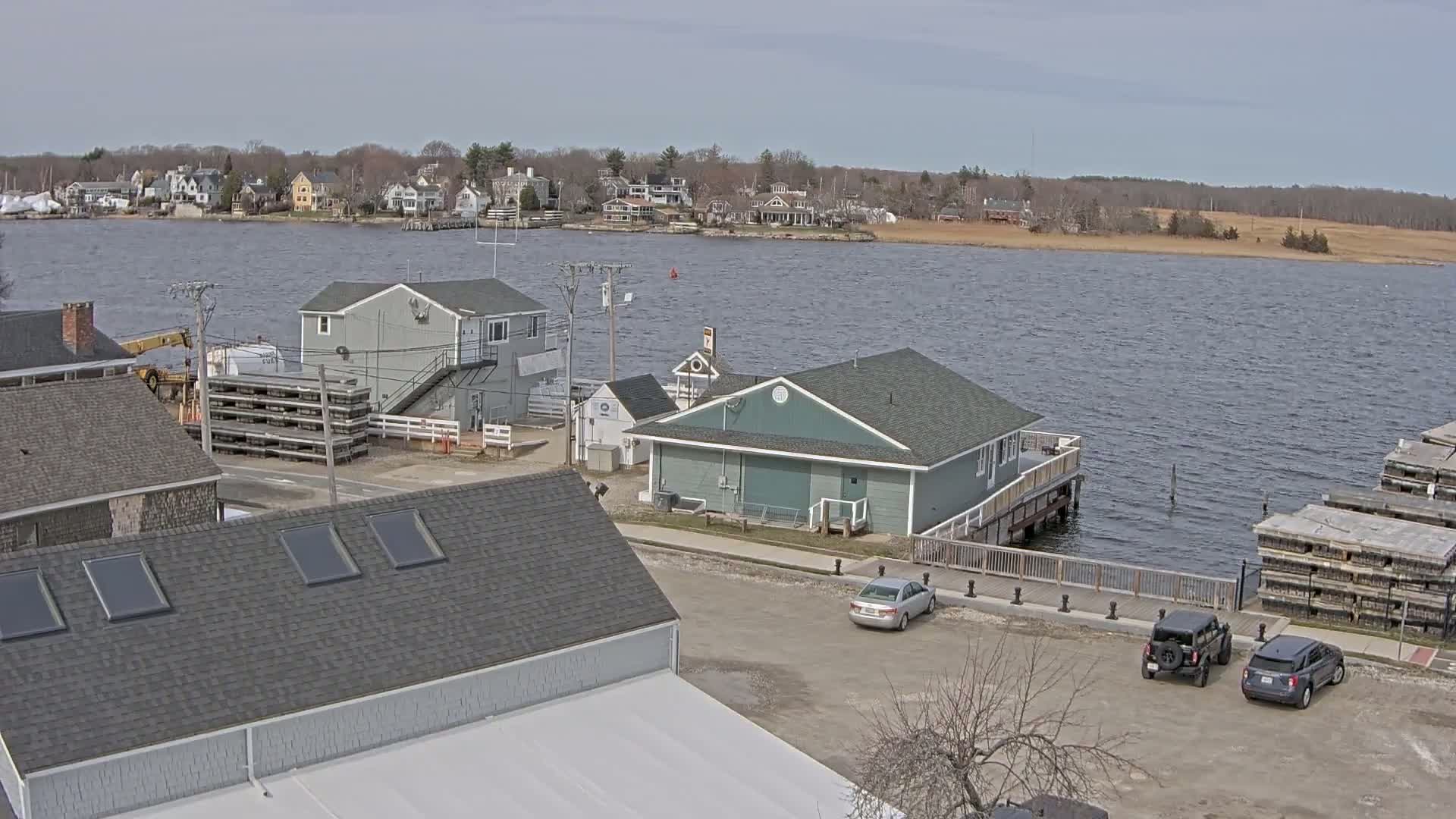 A waterfront view shows several buildings, a parking lot with cars, and stacks of what appear to be wooden pallets near a body of water under a partly cloudy sky.