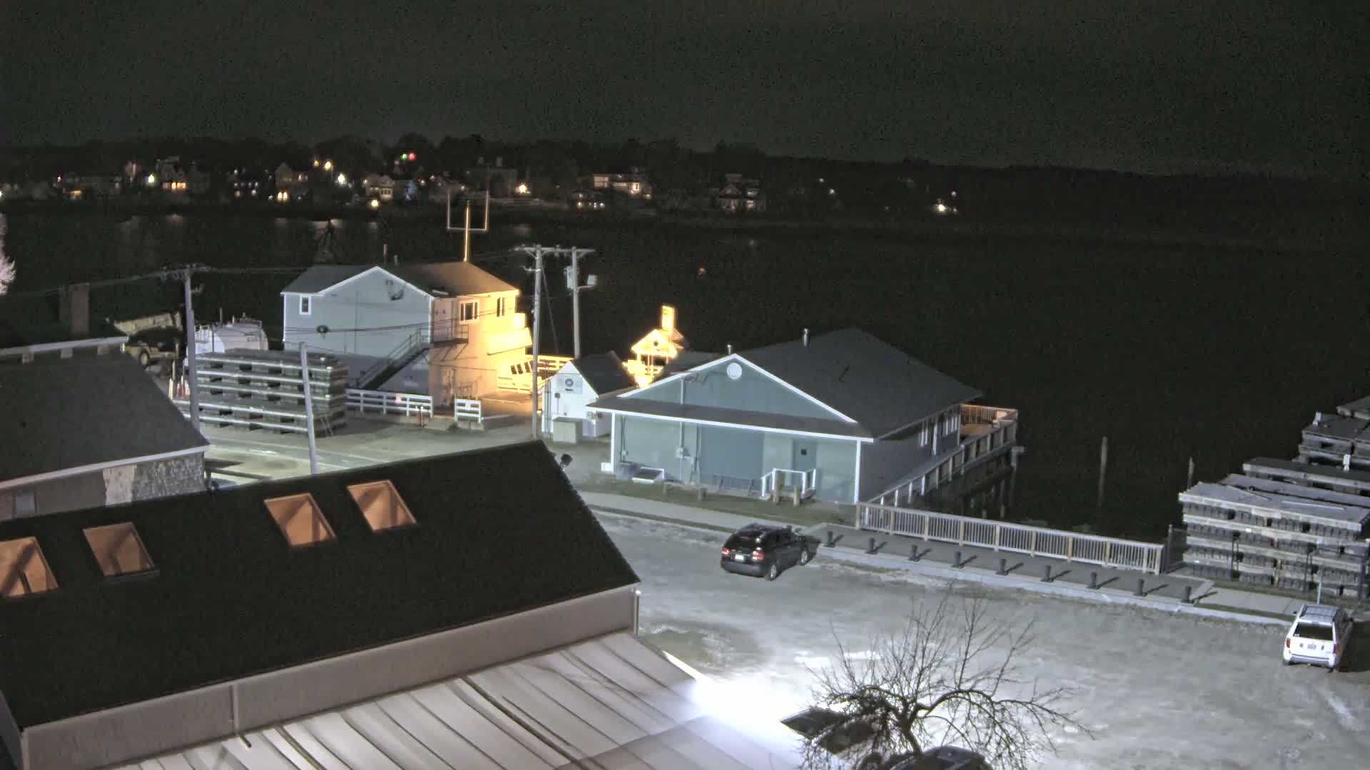 A nighttime, snow-dusted waterfront scene shows several buildings, a few vehicles in a parking lot, and stacks of wooden planks near a body of dark water.