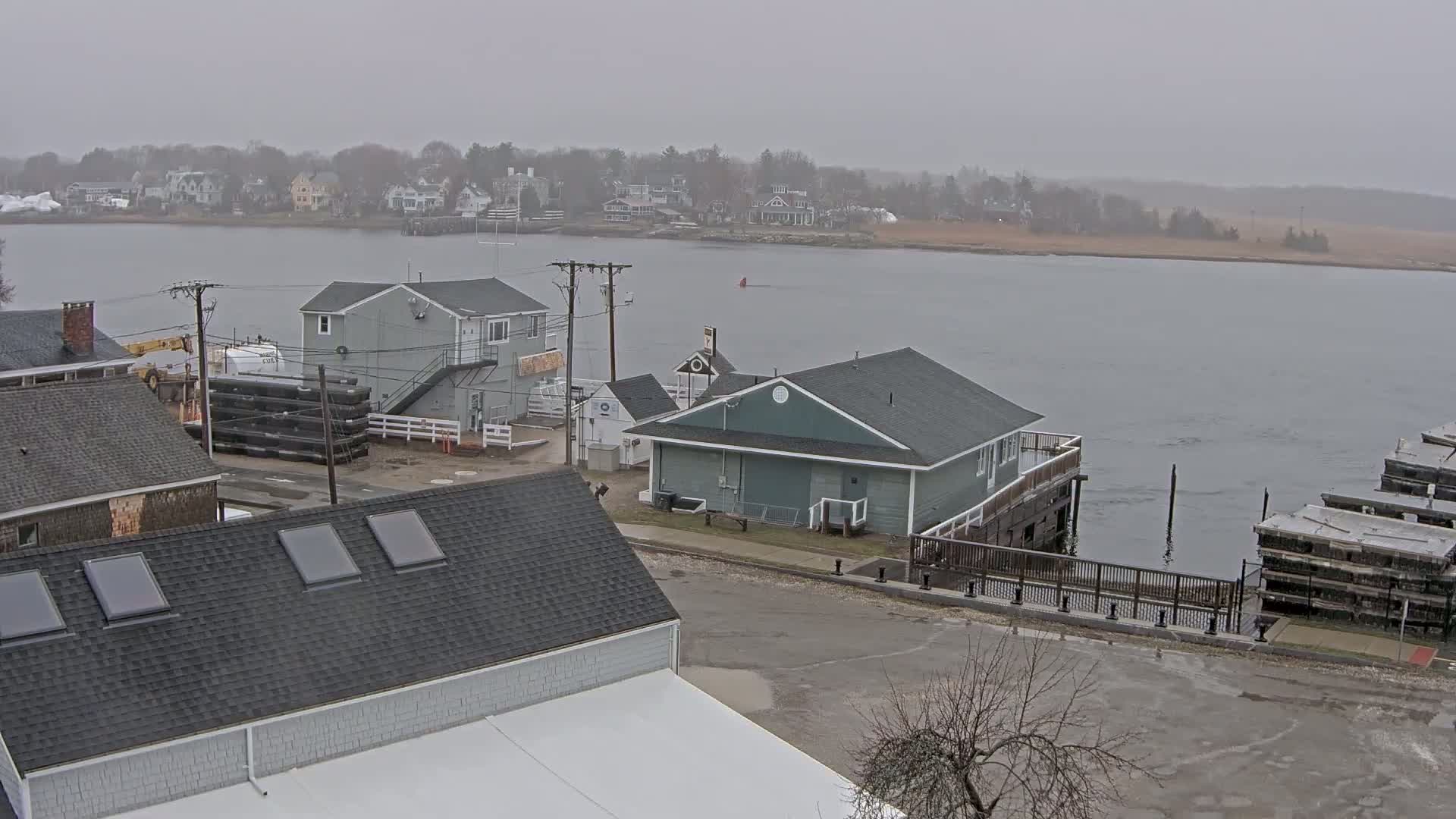 A waterfront scene on an overcast day shows several buildings, a calm river, and a distant shoreline with houses.