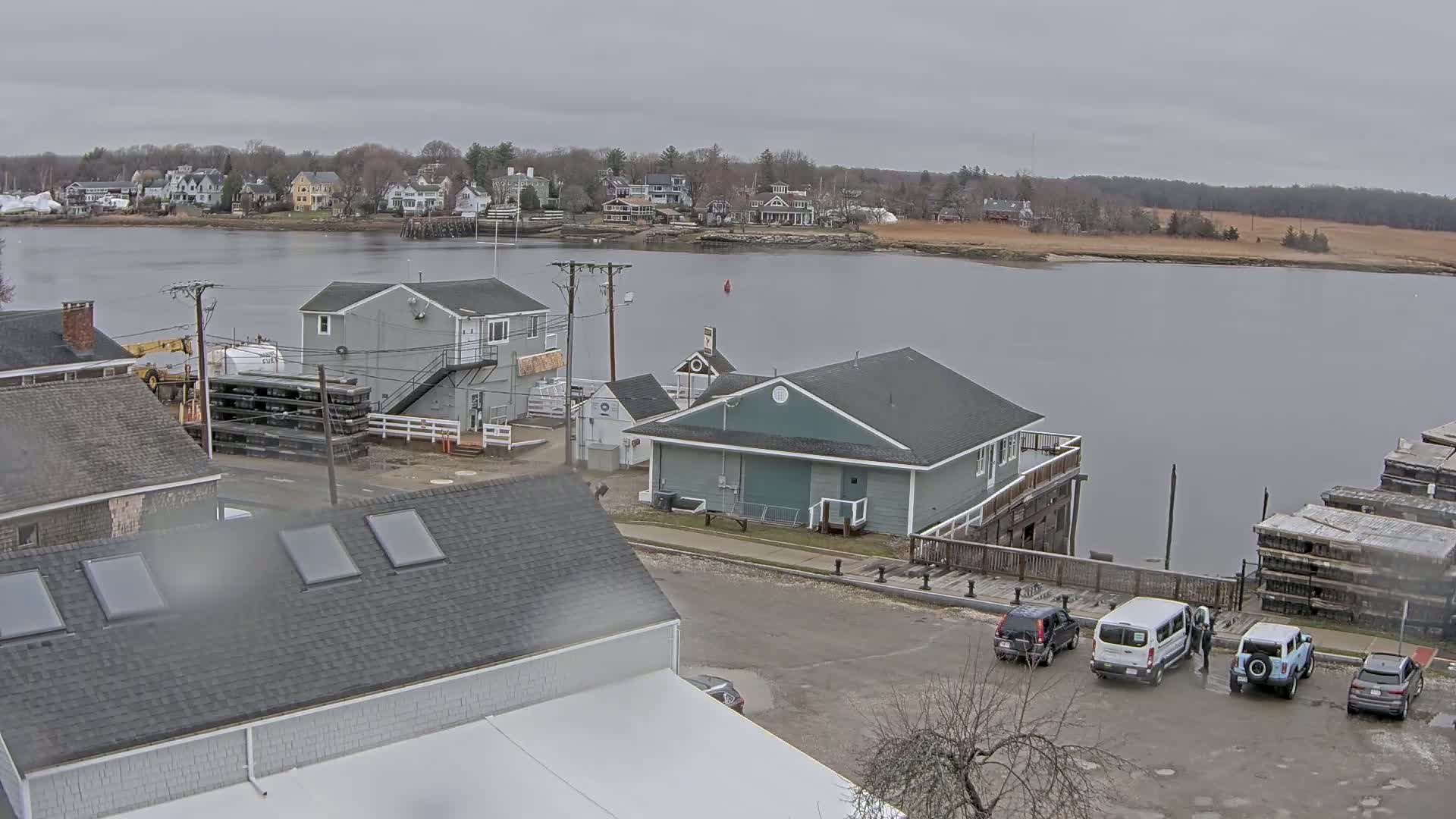 A waterfront scene on an overcast day shows several buildings near a calm river, with cars parked in a lot nearby and a town visible across the water.