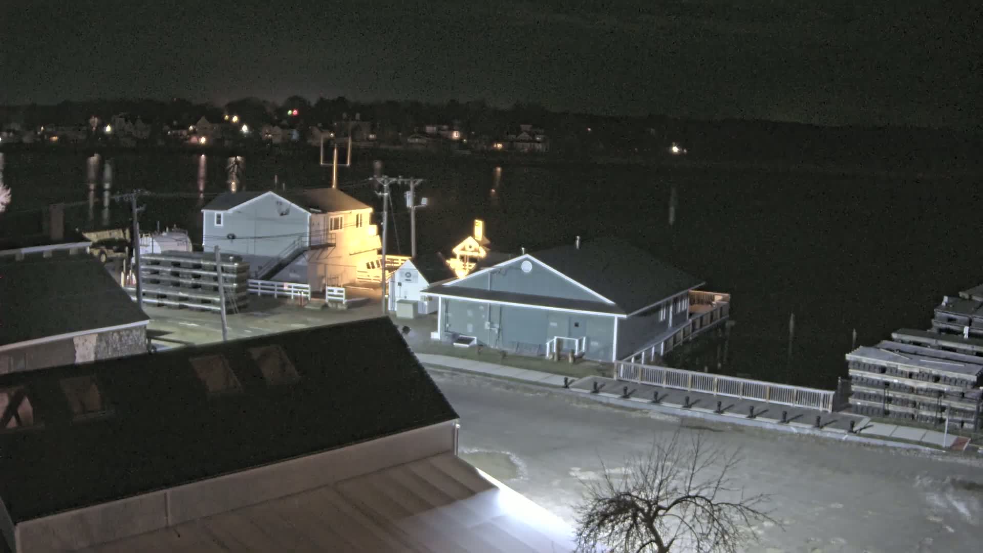 A nighttime view of a waterfront town shows several buildings, a calm body of water reflecting lights from the opposite shore, and stacks of what appear to be wooden planks.