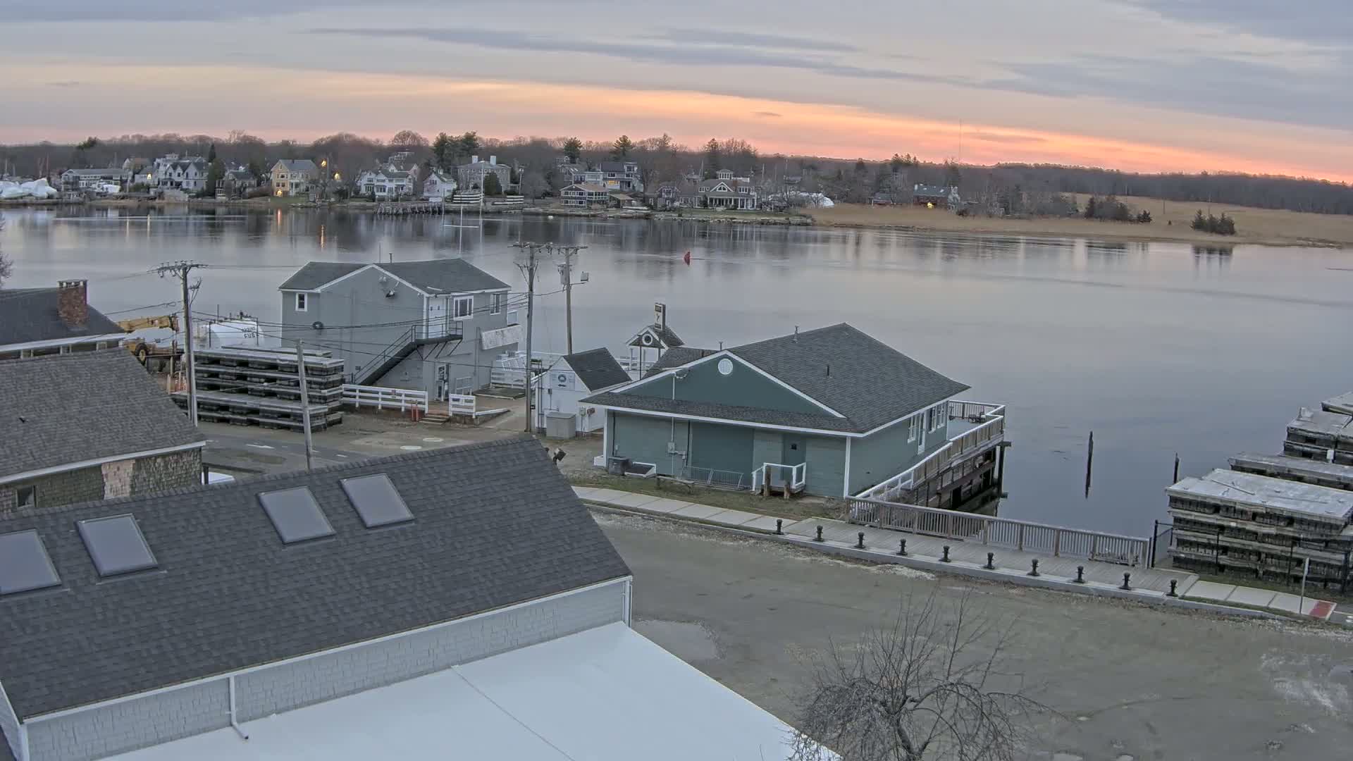 A waterfront scene at dawn shows several buildings near a calm river with a town in the background under a partly cloudy sky.