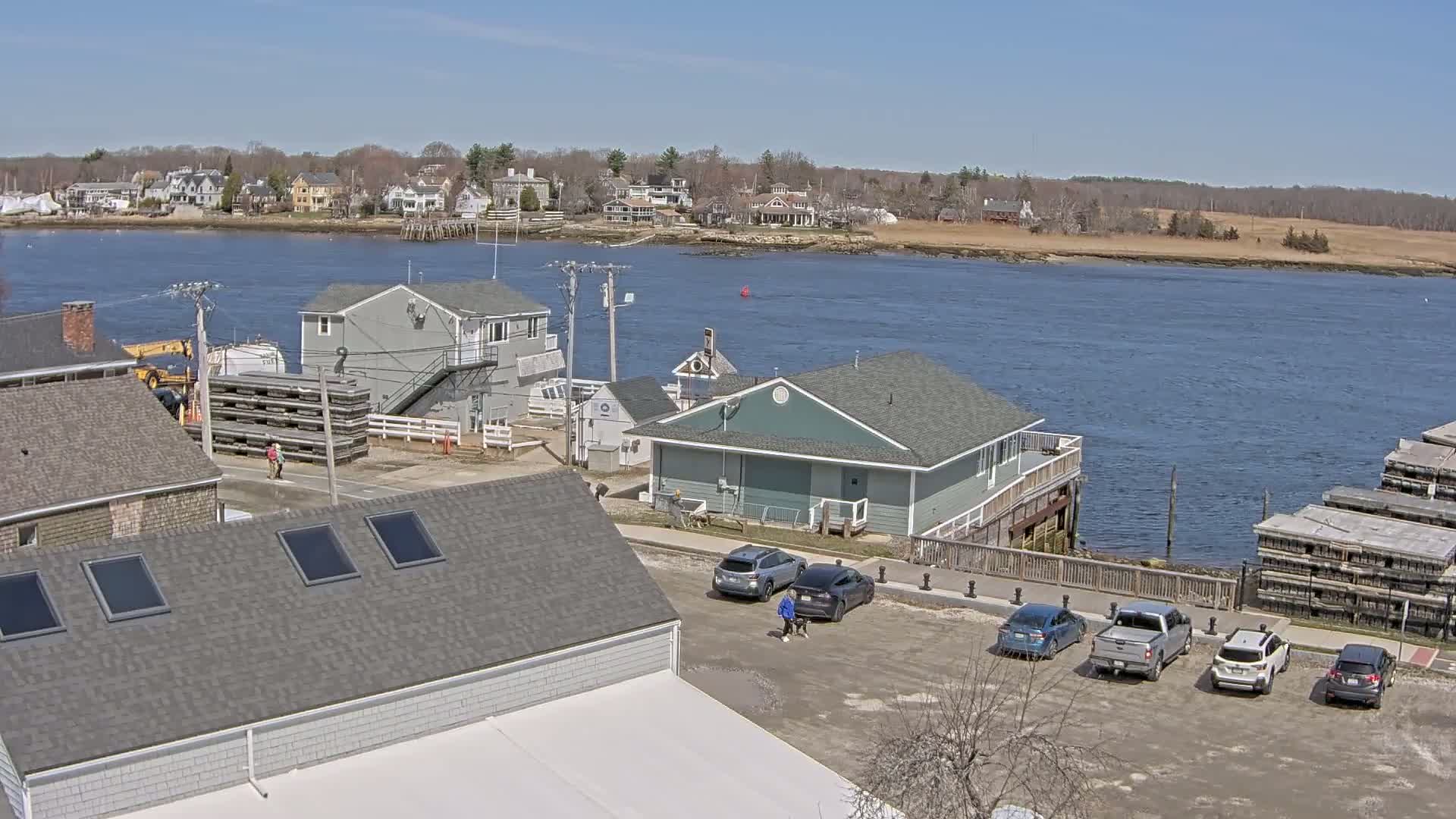 A sunny day overlooking a waterfront town with several houses, a parking lot with cars, and a body of water with stacks of what appear to be oyster cages.
