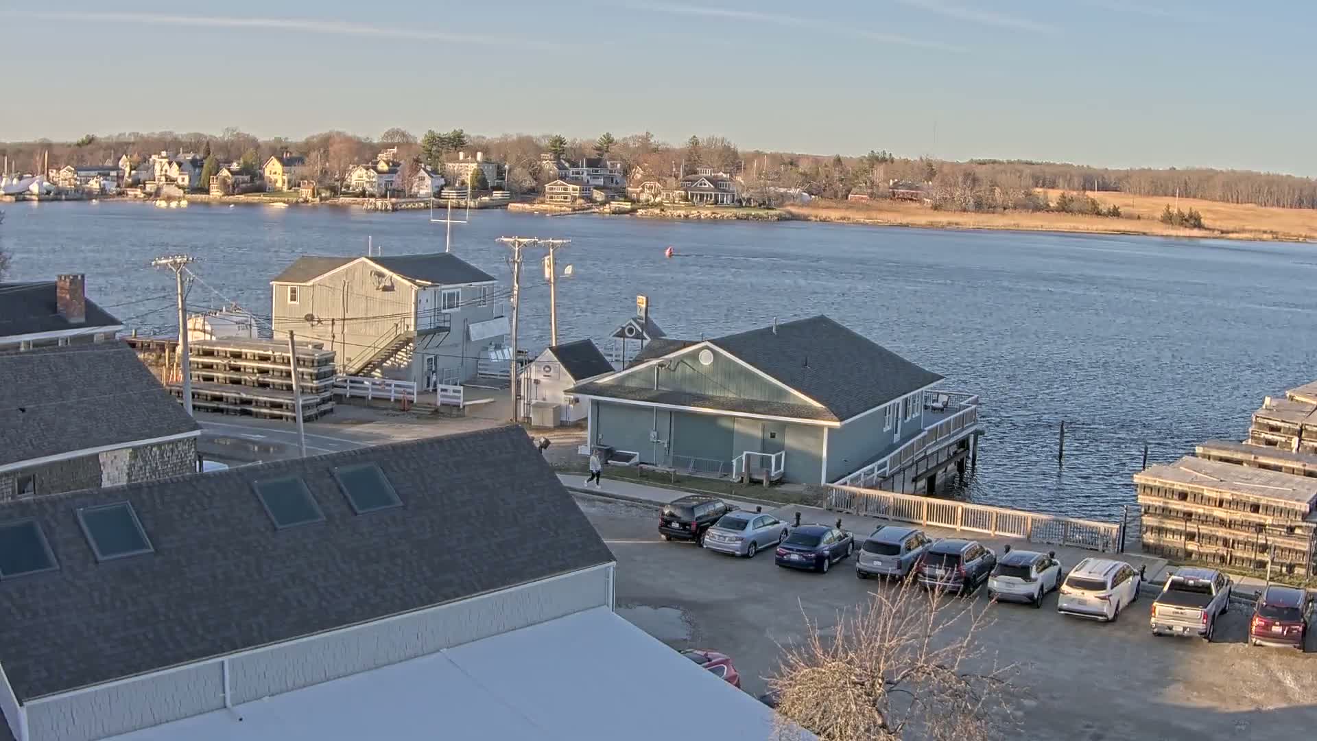 A waterfront town with several buildings, cars in a parking lot, and stacks of what appear to be lobster traps, under a clear, sunny sky.